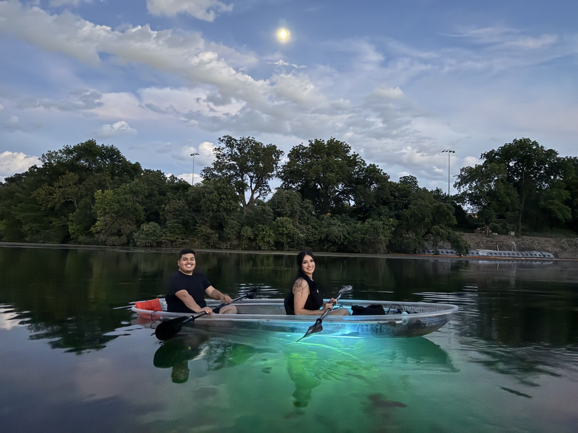 Two people kayaking in a clear kayak on a lake at dusk.  Green lights illuminate below, with trees in the background.