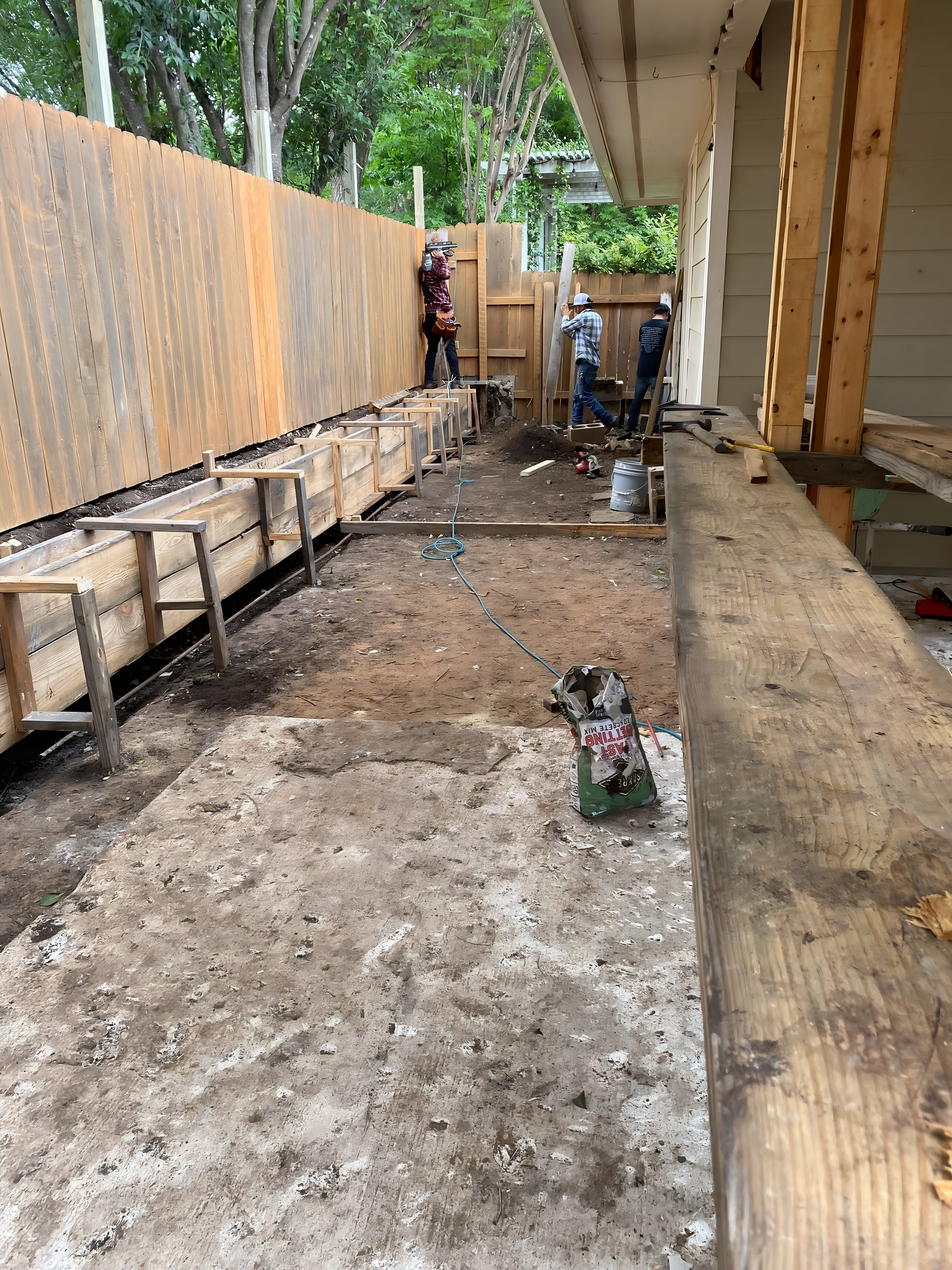 Construction workers at a house, building a concrete form alongside a wooden fence.