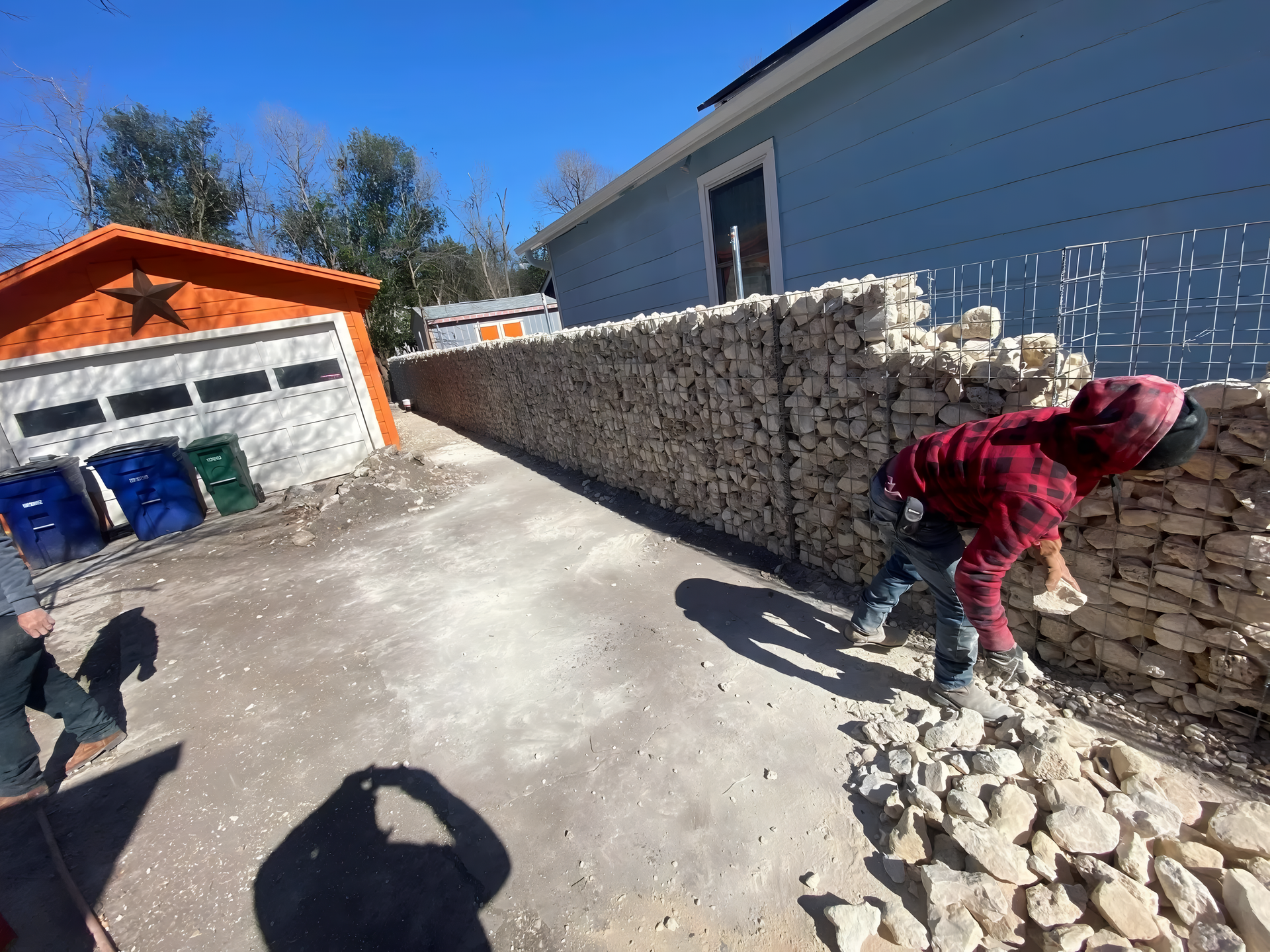 Workers building a stone wall along a blue house in an outdoor setting. One man in a red hoodie is leaning.