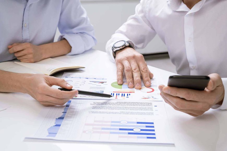 Two people reviewing charts and graphs, one holding a phone, one pointing with a pen.