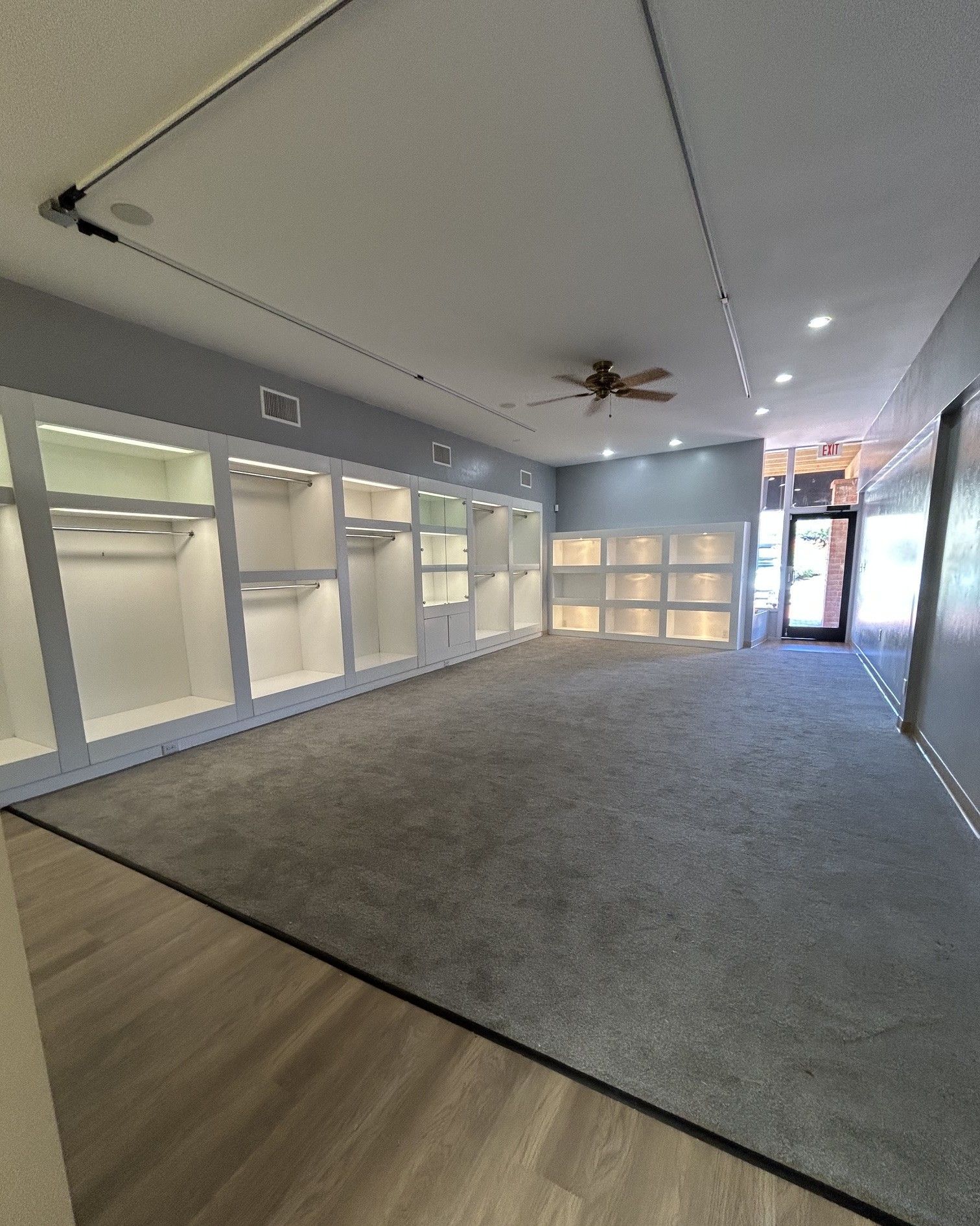 Empty carpeted office lobby with glass-walled rooms and a ceiling fan, viewed from a wood floor entry.