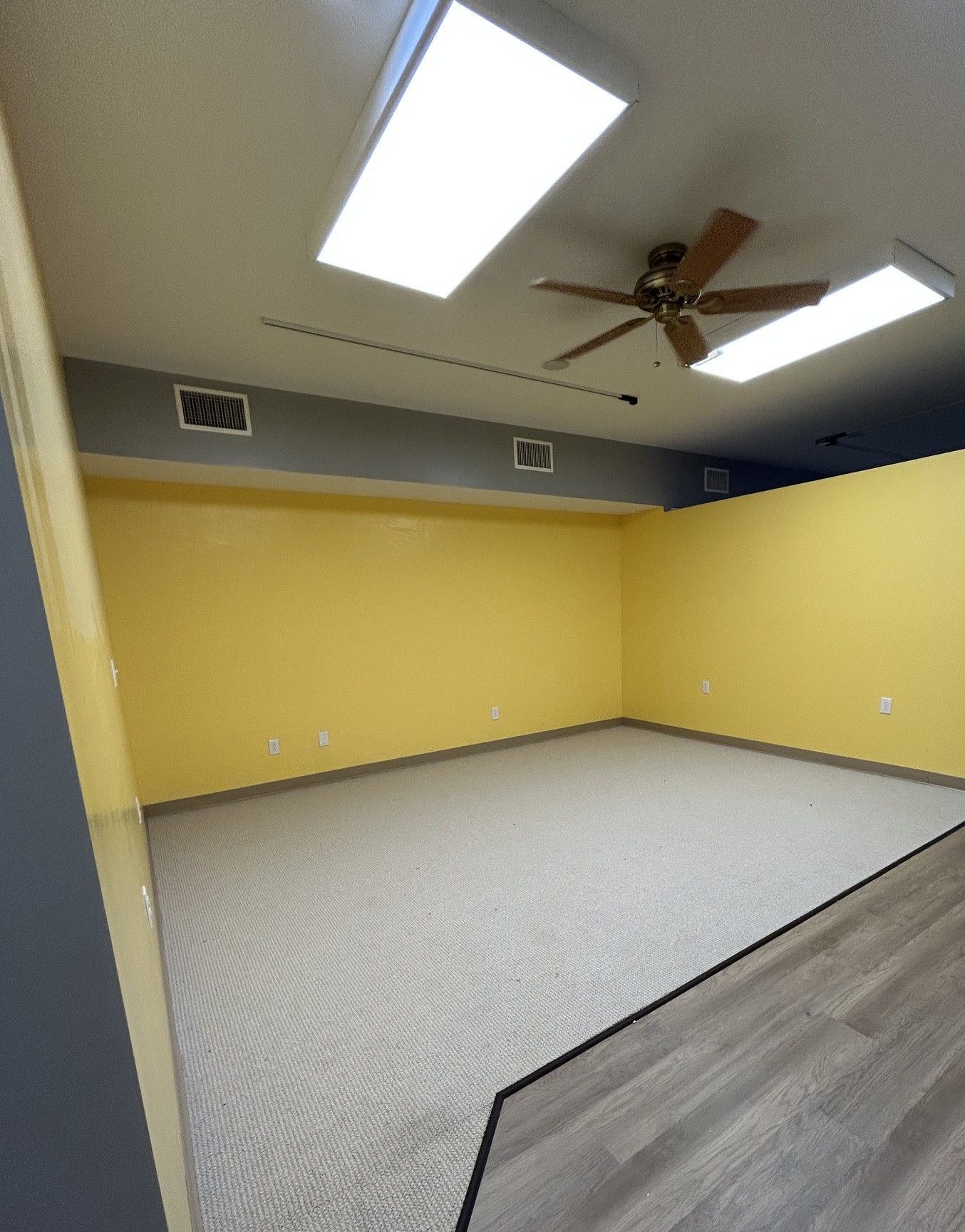 Empty room with yellow walls, beige carpet, fluorescent lights, and a ceiling fan.