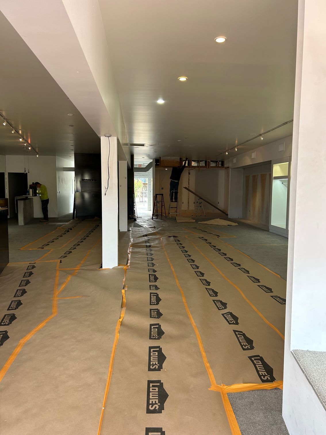 Empty indoor room under renovation, with white columns, exposed floor protection, and recessed ceiling lights.