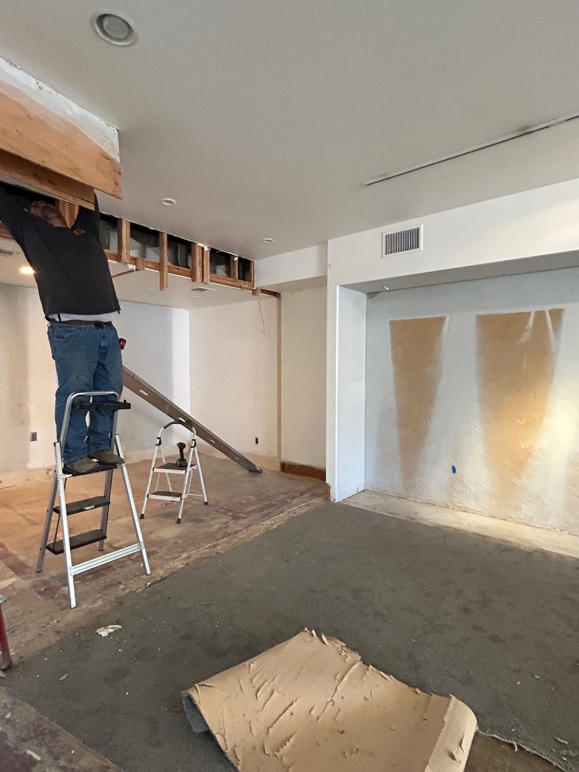 Worker on a ladder installing ceiling drywall in an unfinished room with exposed framing.