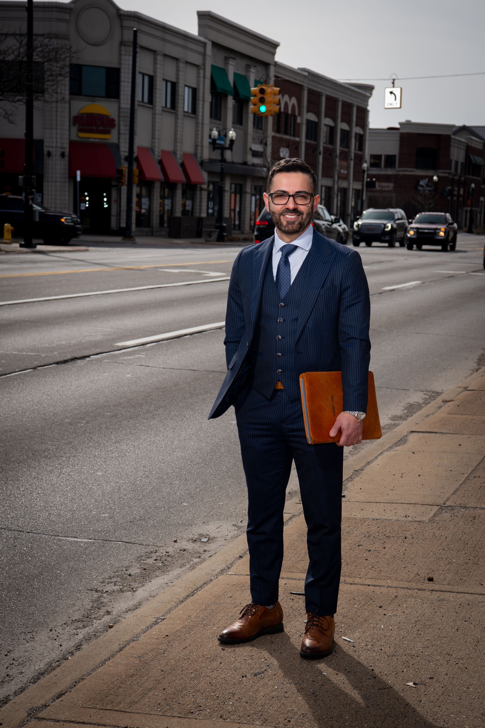 Man in a navy suit holding a brown portfolio, standing on a city sidewalk beside an empty street.