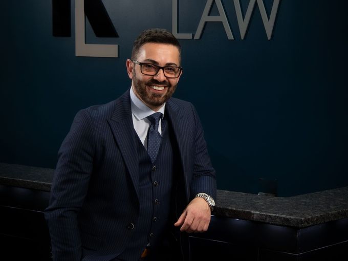 Man in a dark suit leaning on a marble counter in a law office, smiling at the camera.