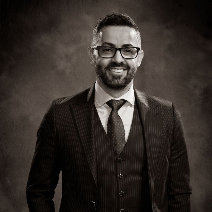 Smiling man in glasses and a suit, posed against a dark studio backdrop.
