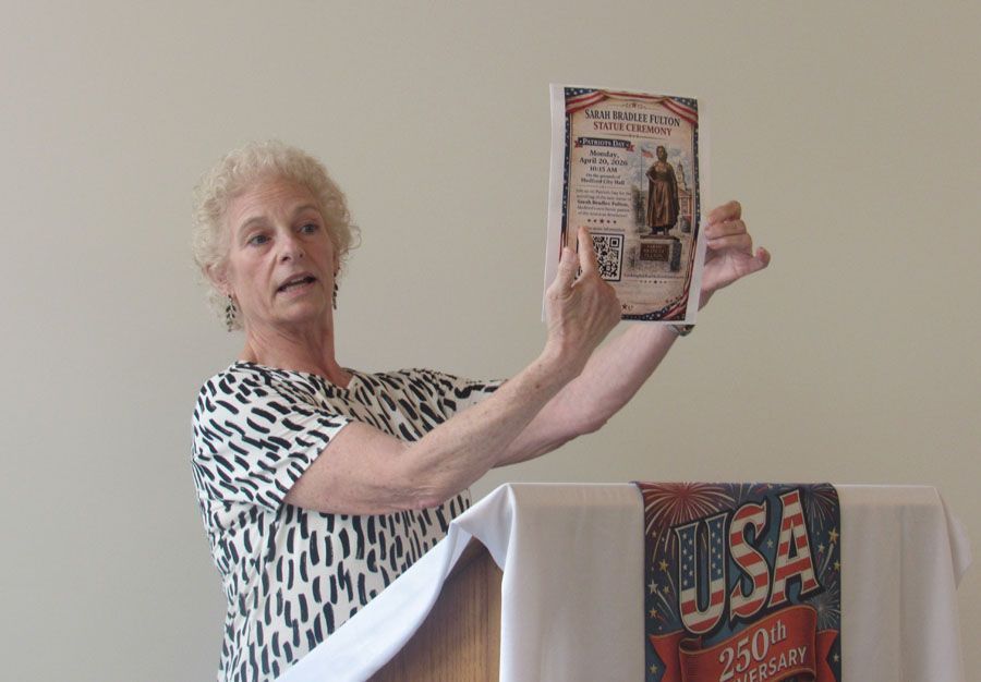 Woman presenting a USA 250th anniversary booklet beside a podium draped with a USA banner