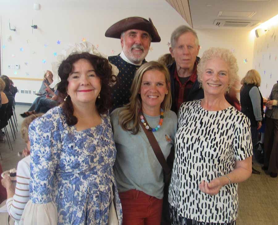 Five smiling adults posing together indoors at a social event, with bright walls and paintings in the background.