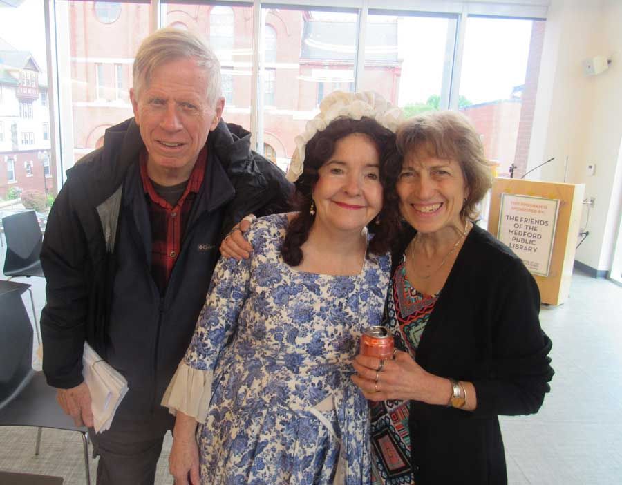 Three people posing together indoors, smiling, with large windows in the background.