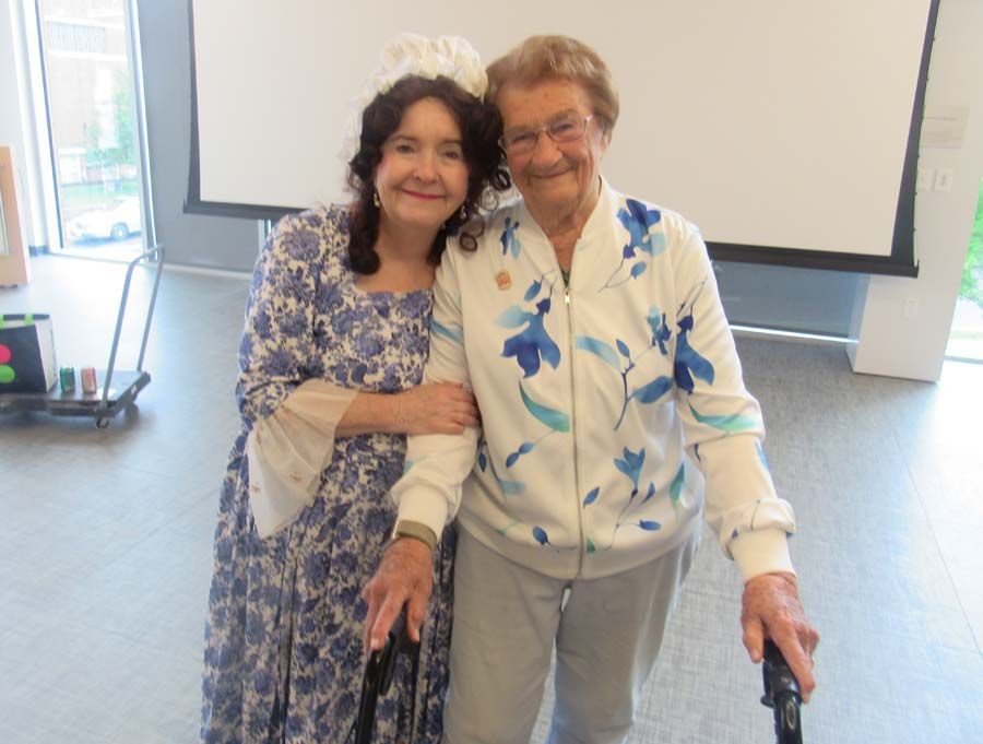 Two people smiling and posing together indoors, one using a walker, in front of a projector screen.