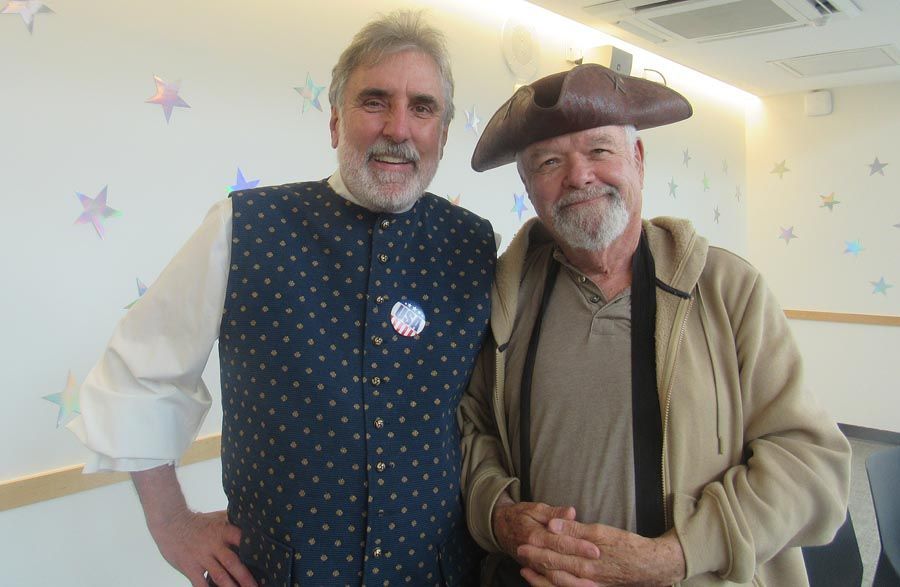 Two men smiling indoors, one in a blue vest and one in a tan jacket and hat