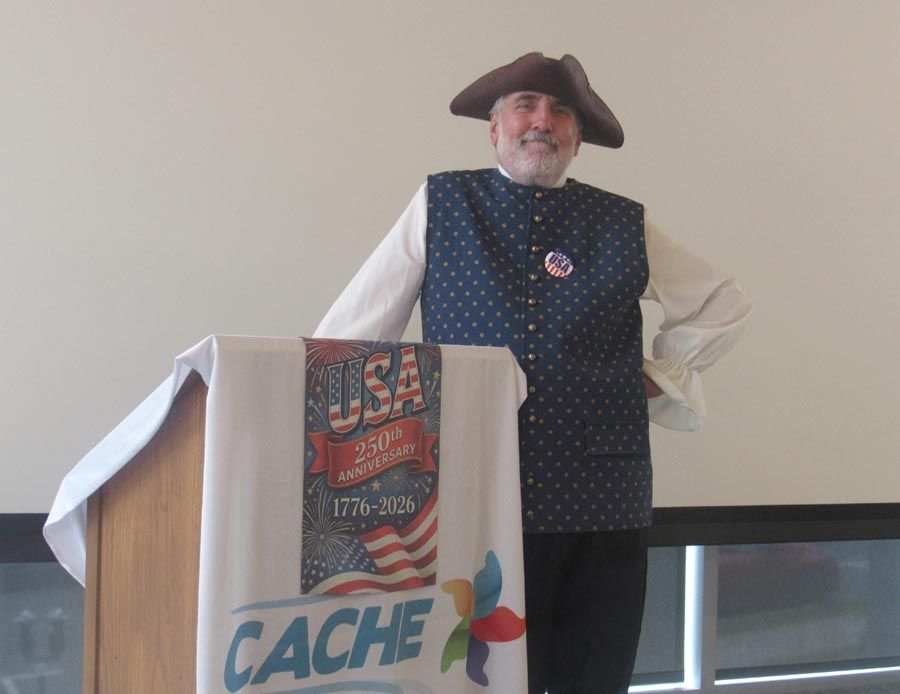 Man in tricorn hat standing at a podium with a USA CACHE banner