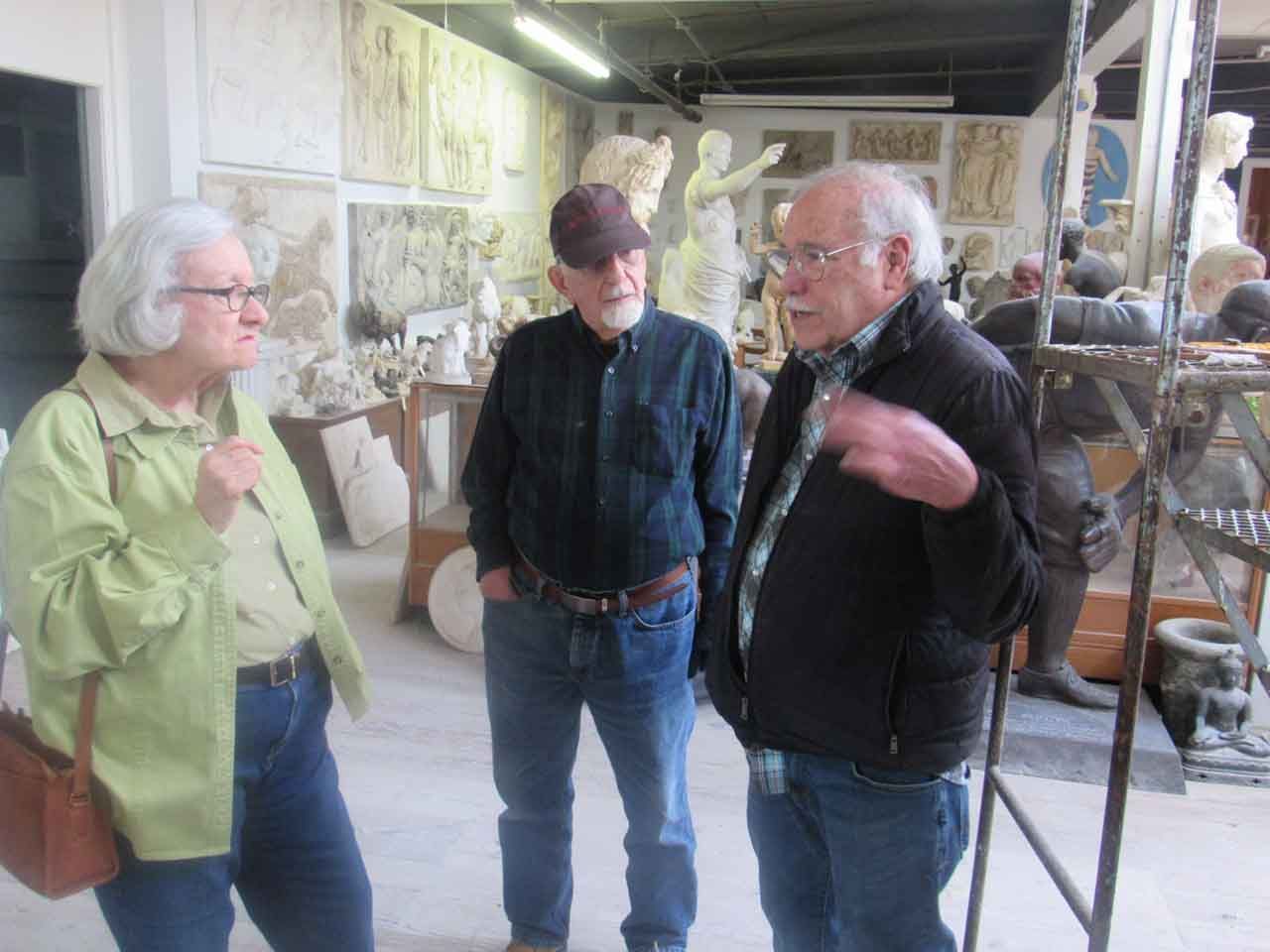 Three people in a studio with sculptures. One gestures while speaking. Walls have art pieces.