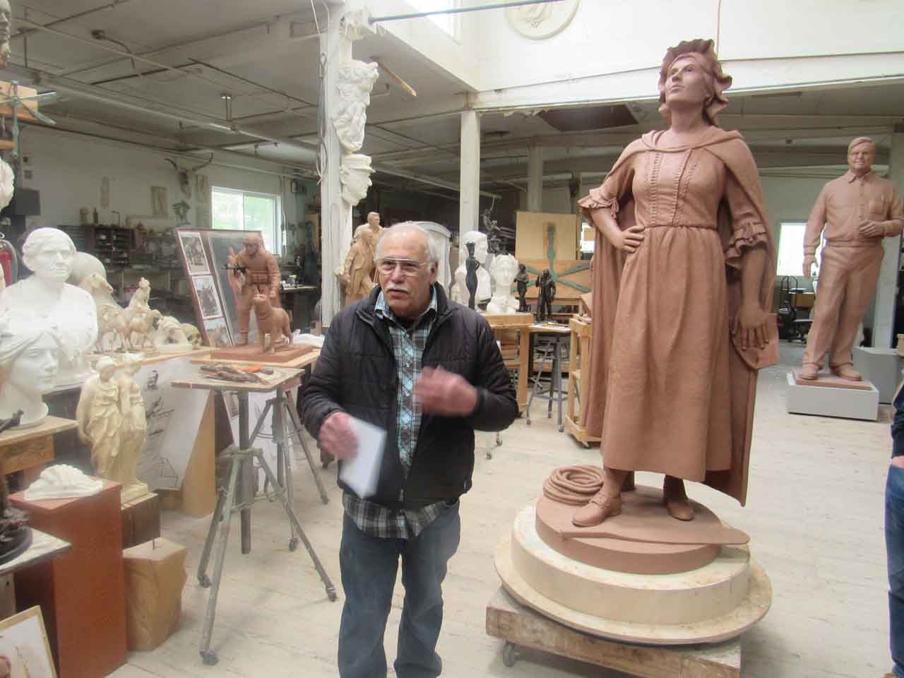 Man standing in a studio with large clay sculptures. A woman figure stands tall, looking upward.