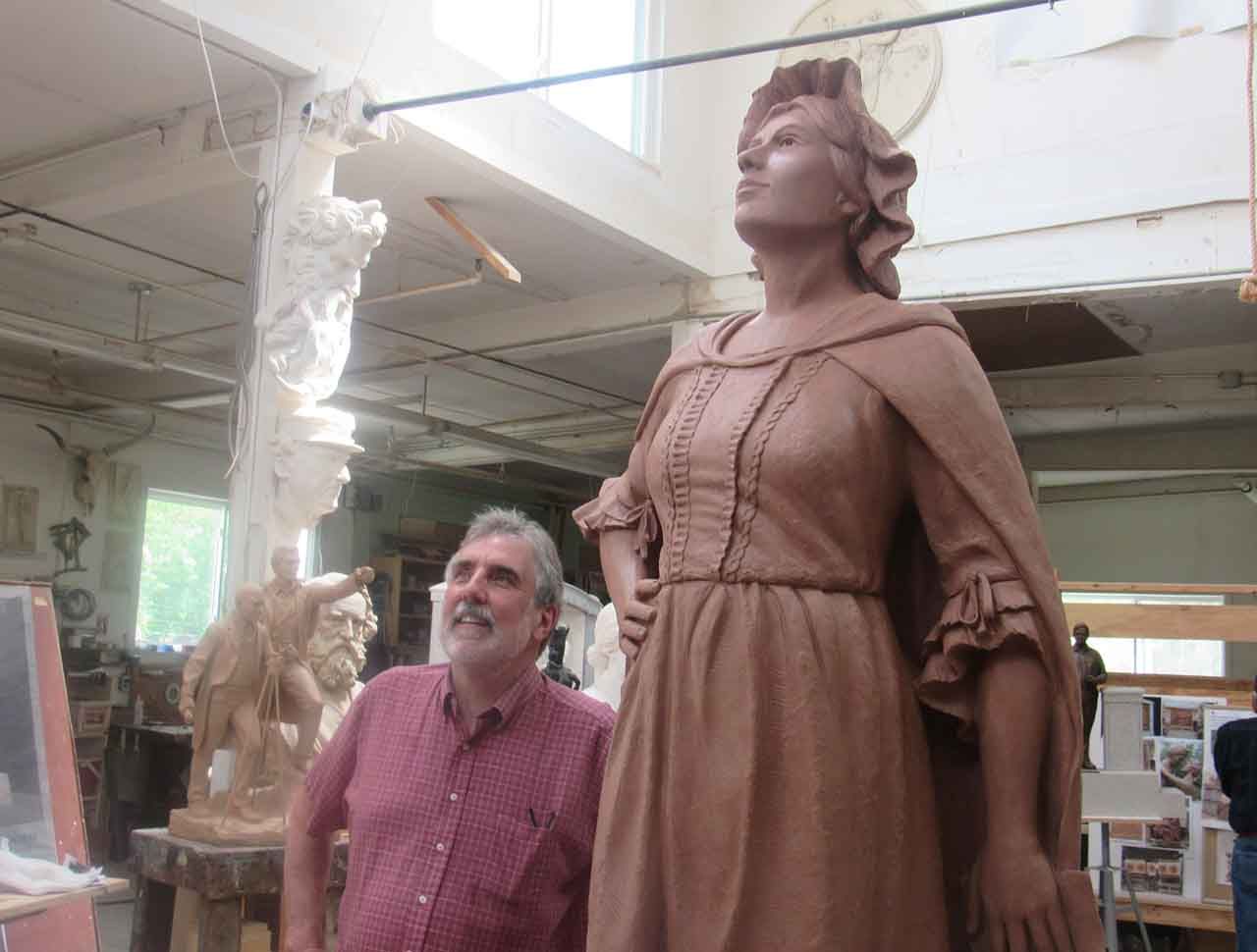 Man looks up at a large clay sculpture of a woman in a studio. The sculpture wears a period dress.