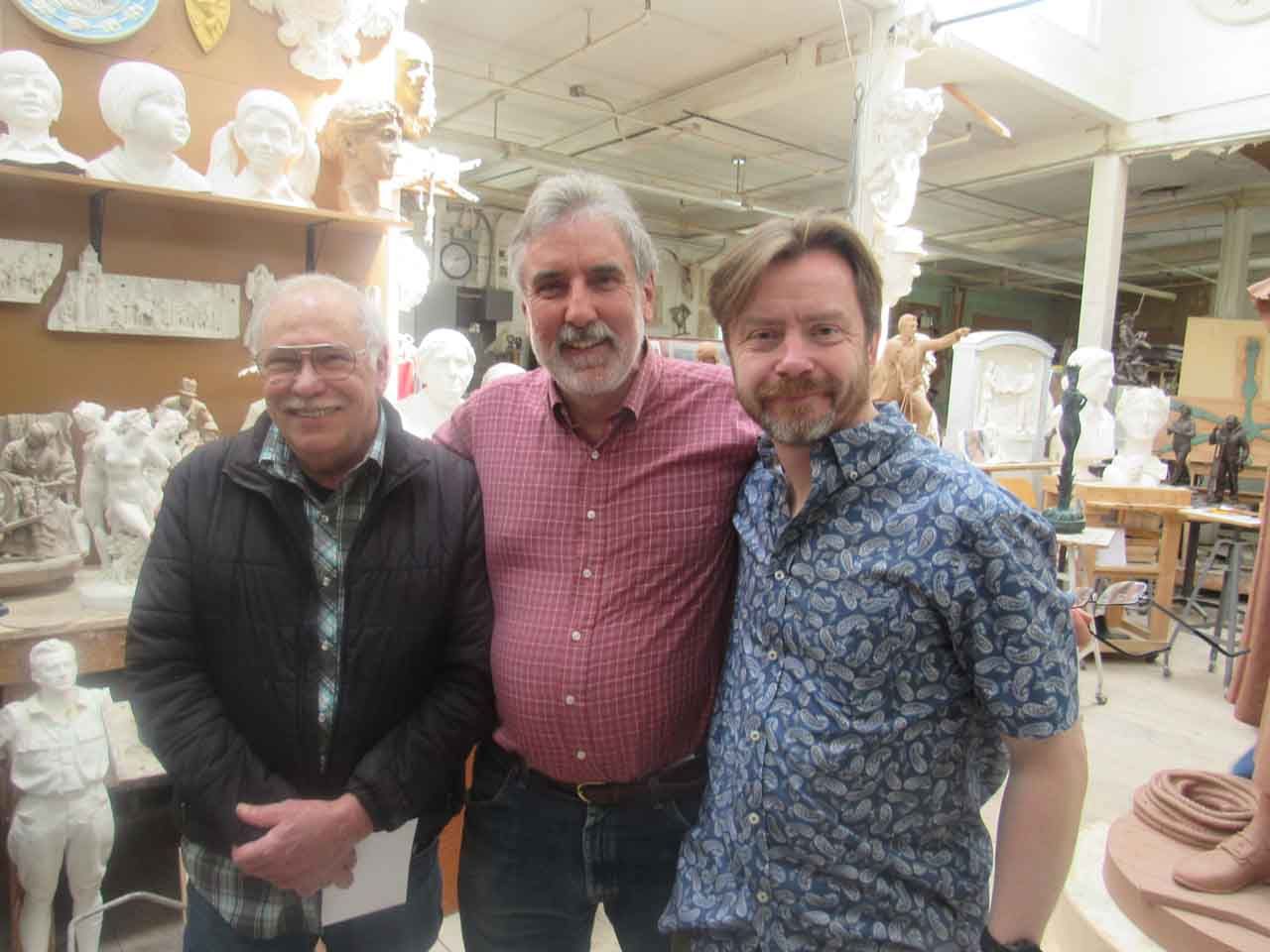 Three men pose for a photo in a sculptor's studio, surrounded by sculptures and busts.