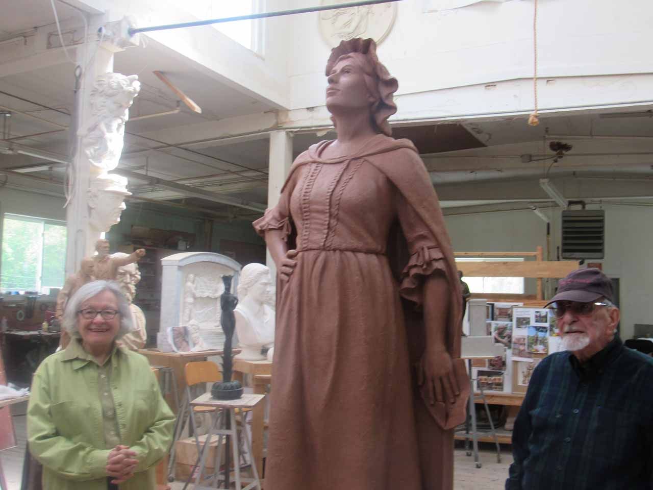 Woman sculpts large statue. Two people pose in front. Workshop. Statue is clay-colored.
