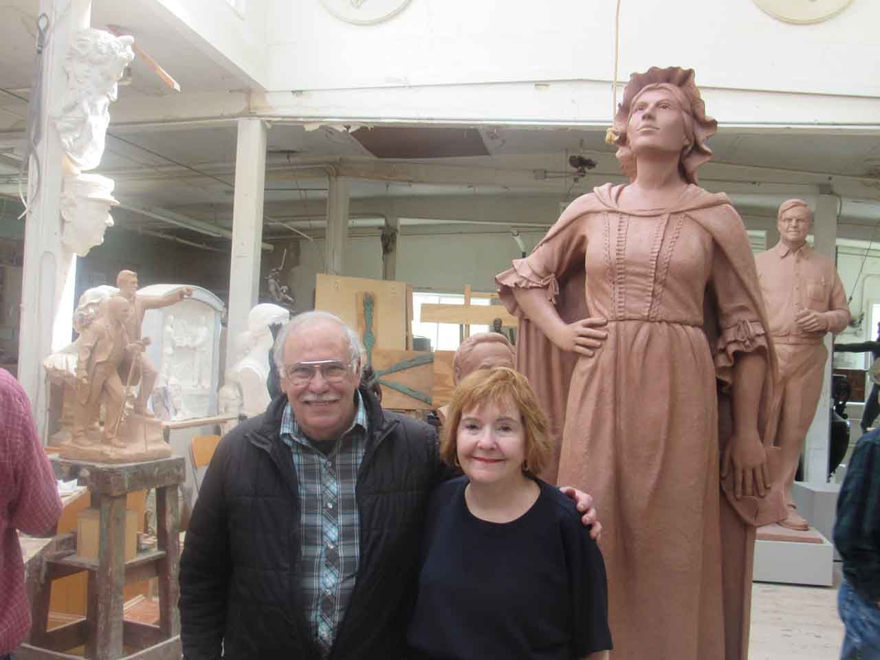 Man and woman pose next to a large clay statue of a woman in a workshop.
