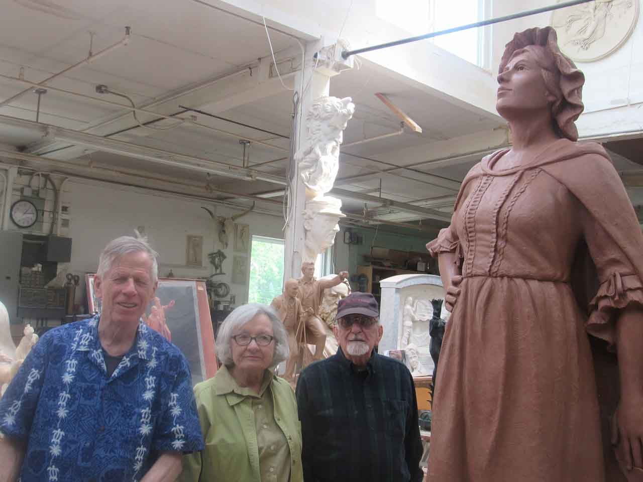 Four people stand near a large clay sculpture of a woman in a studio. The woman gazes upwards.