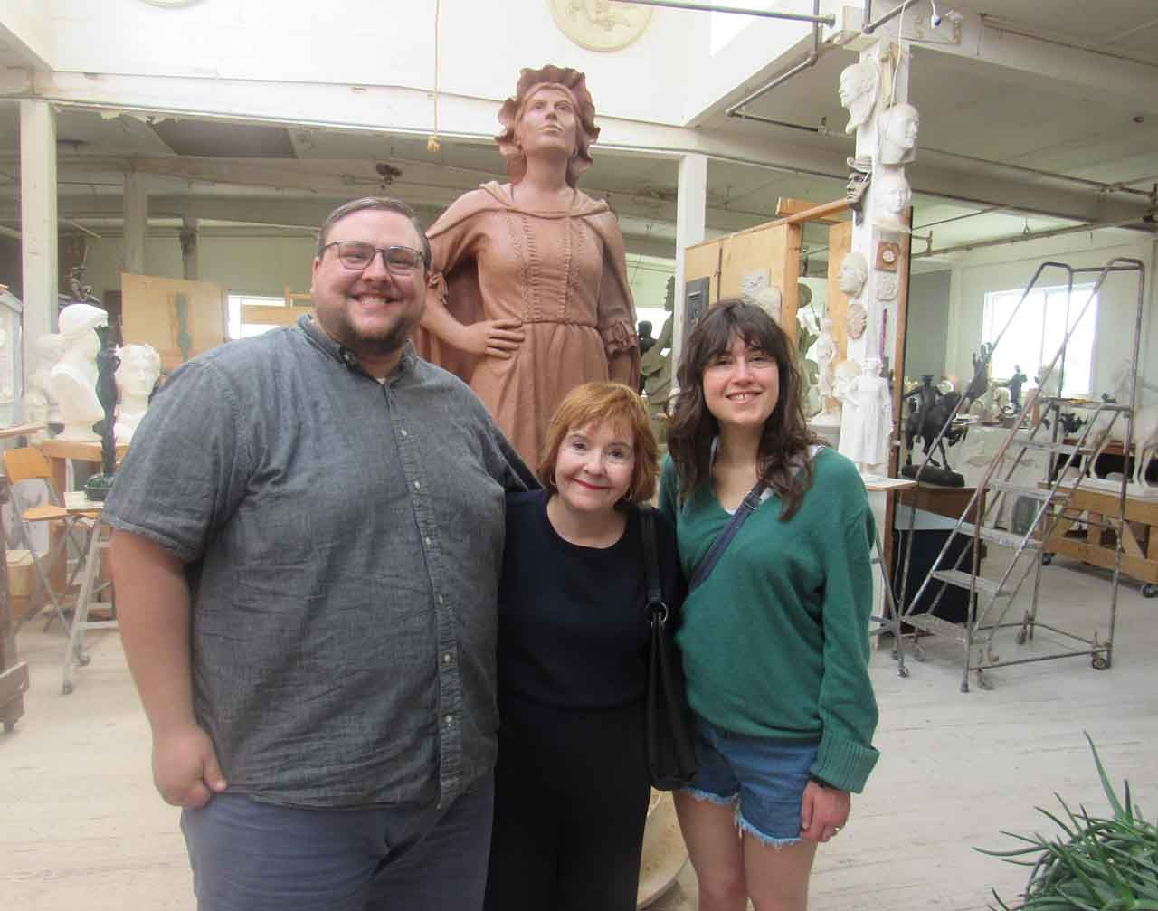 Three people pose in front of a statue in a studio. The statue is a figure in a flowing gown.