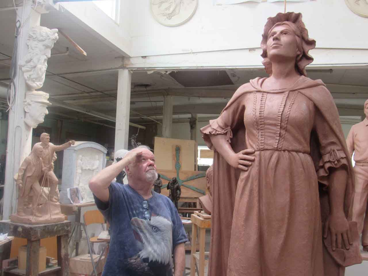 Sculptor beside a large clay statue of a woman in a workshop; statue has cloak, hand on hip, looking up.
