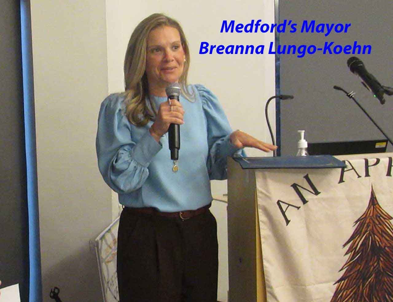 Medford's Mayor Breanna Lungo-Koehn speaking at a podium, light blue blouse, holding a microphone.