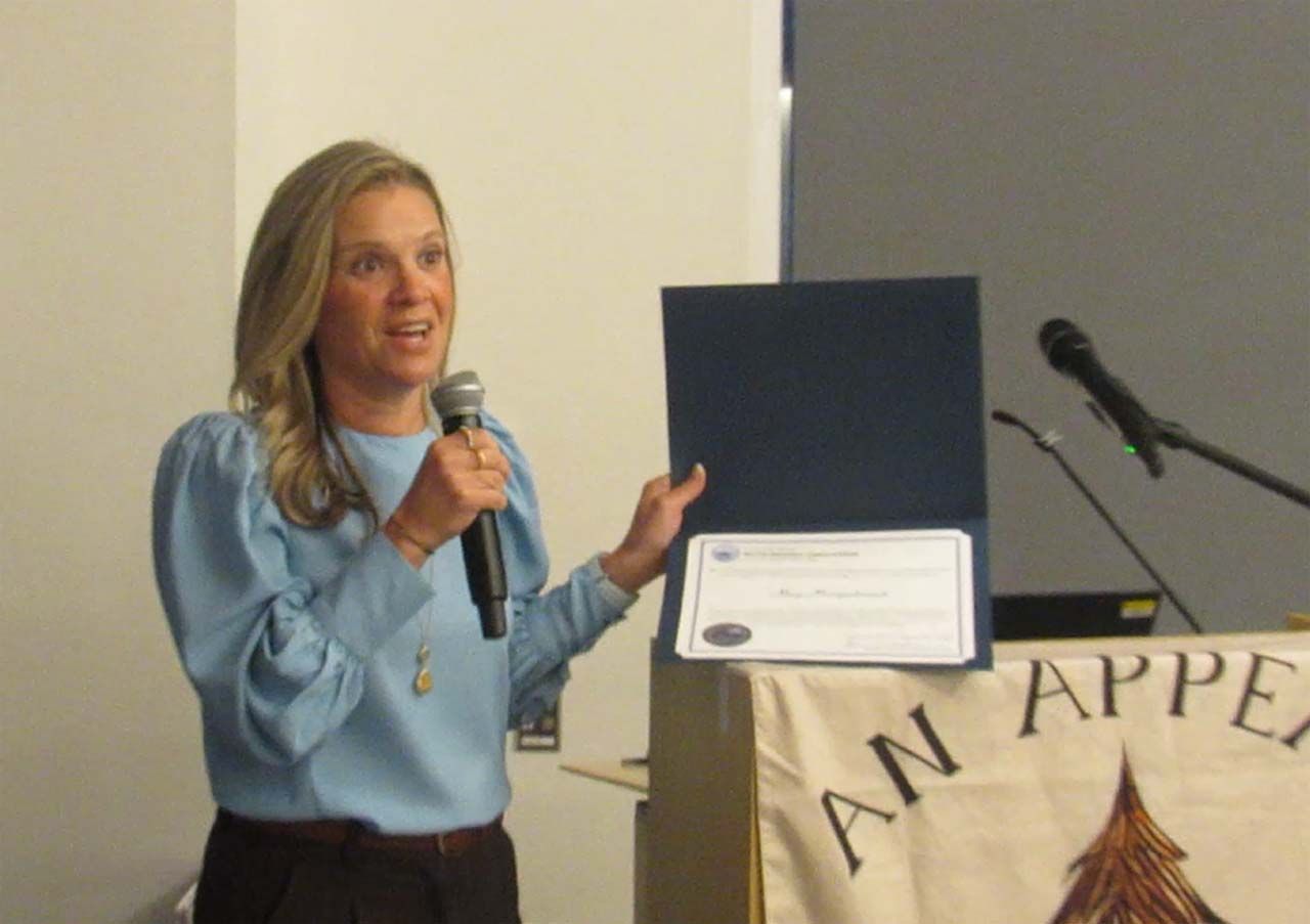 Woman in blue top, holding a microphone and document, giving a presentation at a podium.