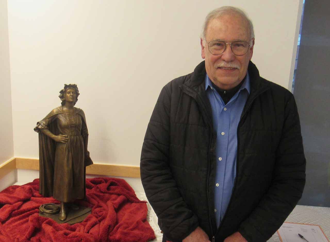 Man stands next to a bronze statue on a red cloth. Statue depicts a woman. White wall.