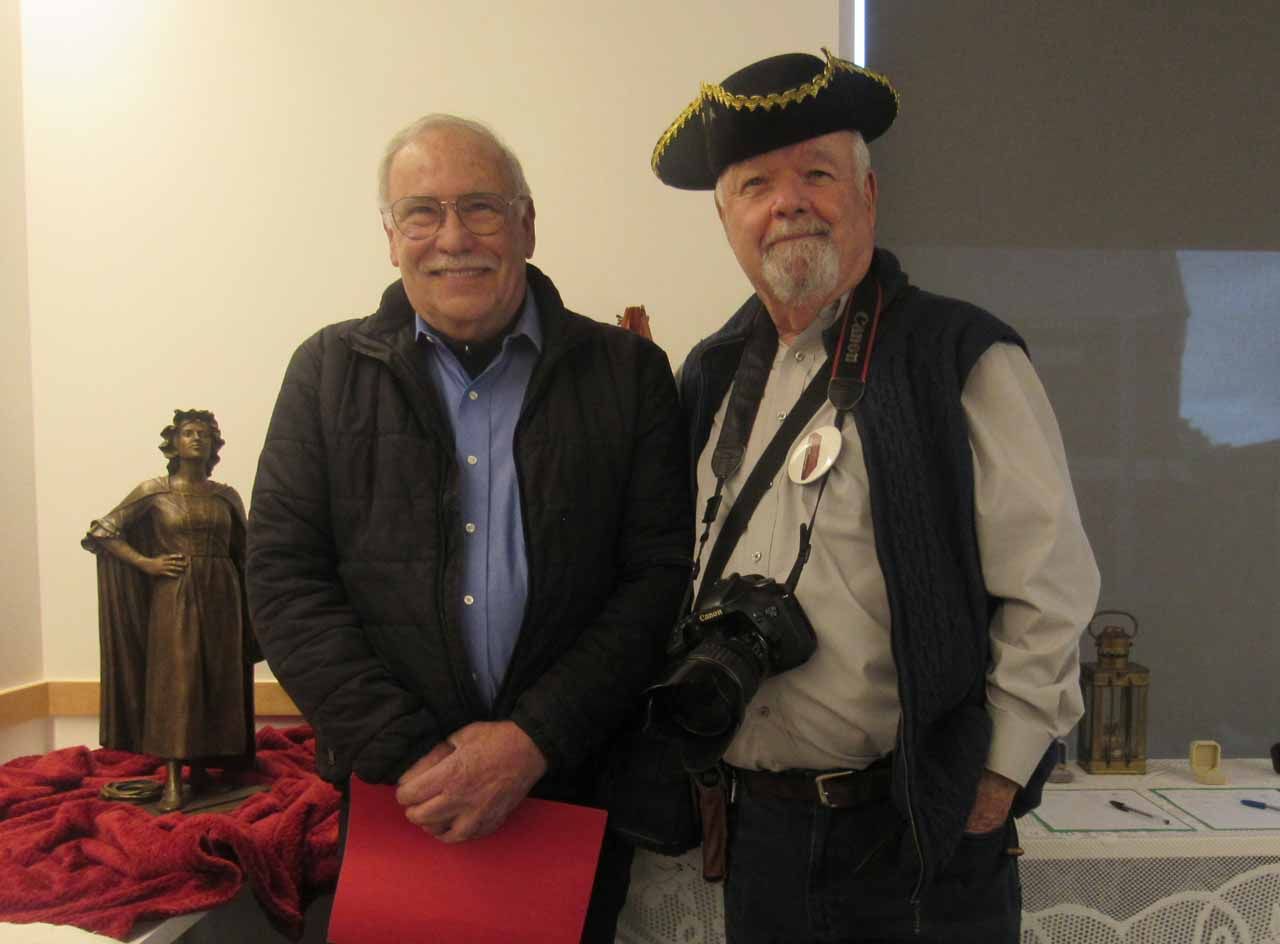 Two men pose near a statue. One wears a pirate hat and holds a camera. A red paper is in the other man's hand.