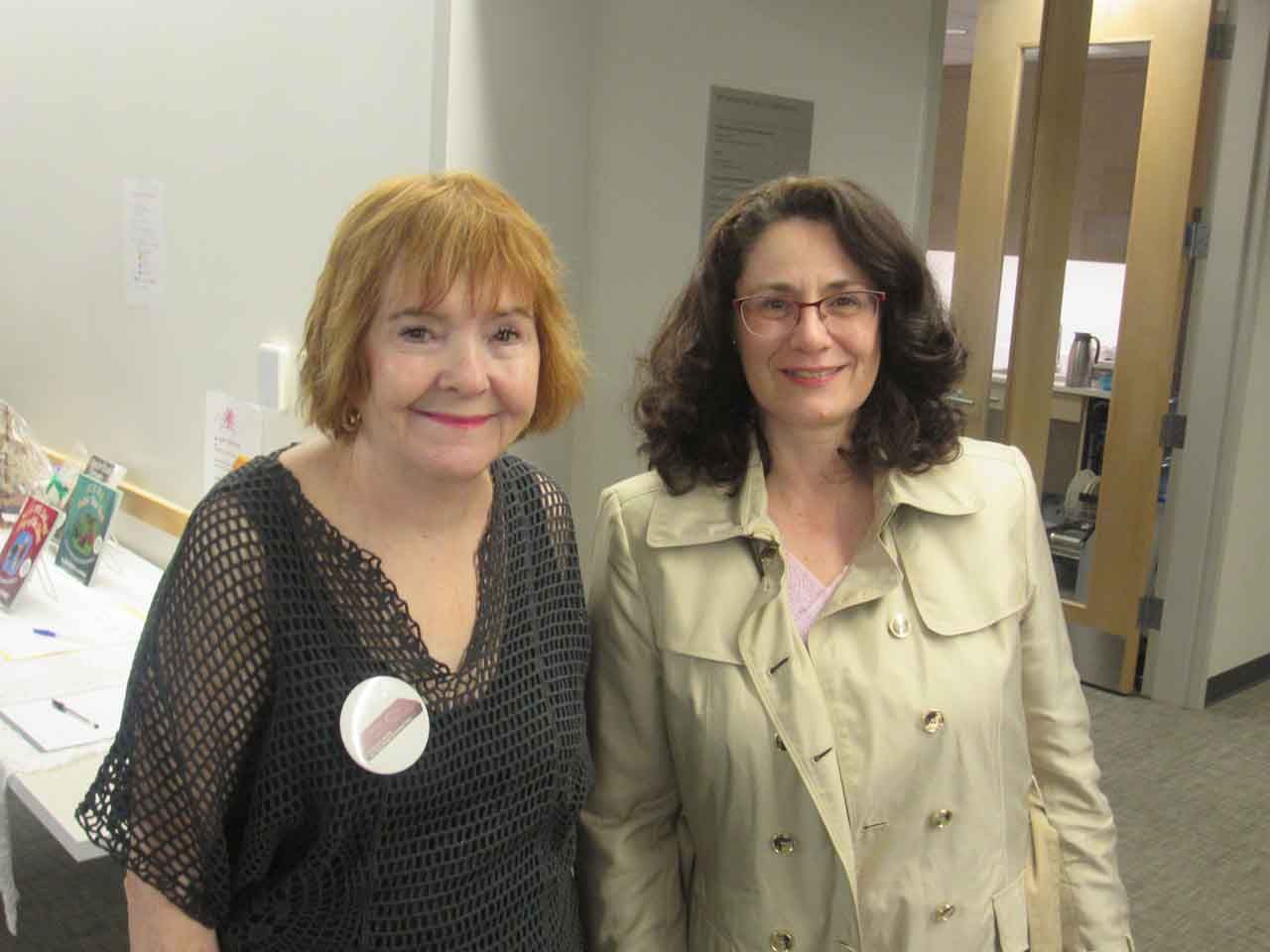 Two women smiling, one in a black top with a pin, the other in a tan coat. Indoor setting, white walls.
