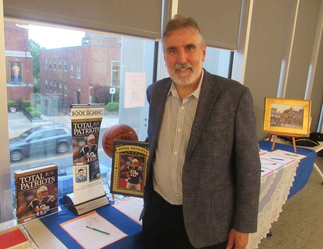 Man in a blazer stands by table with sports memorabilia and books, near a window.