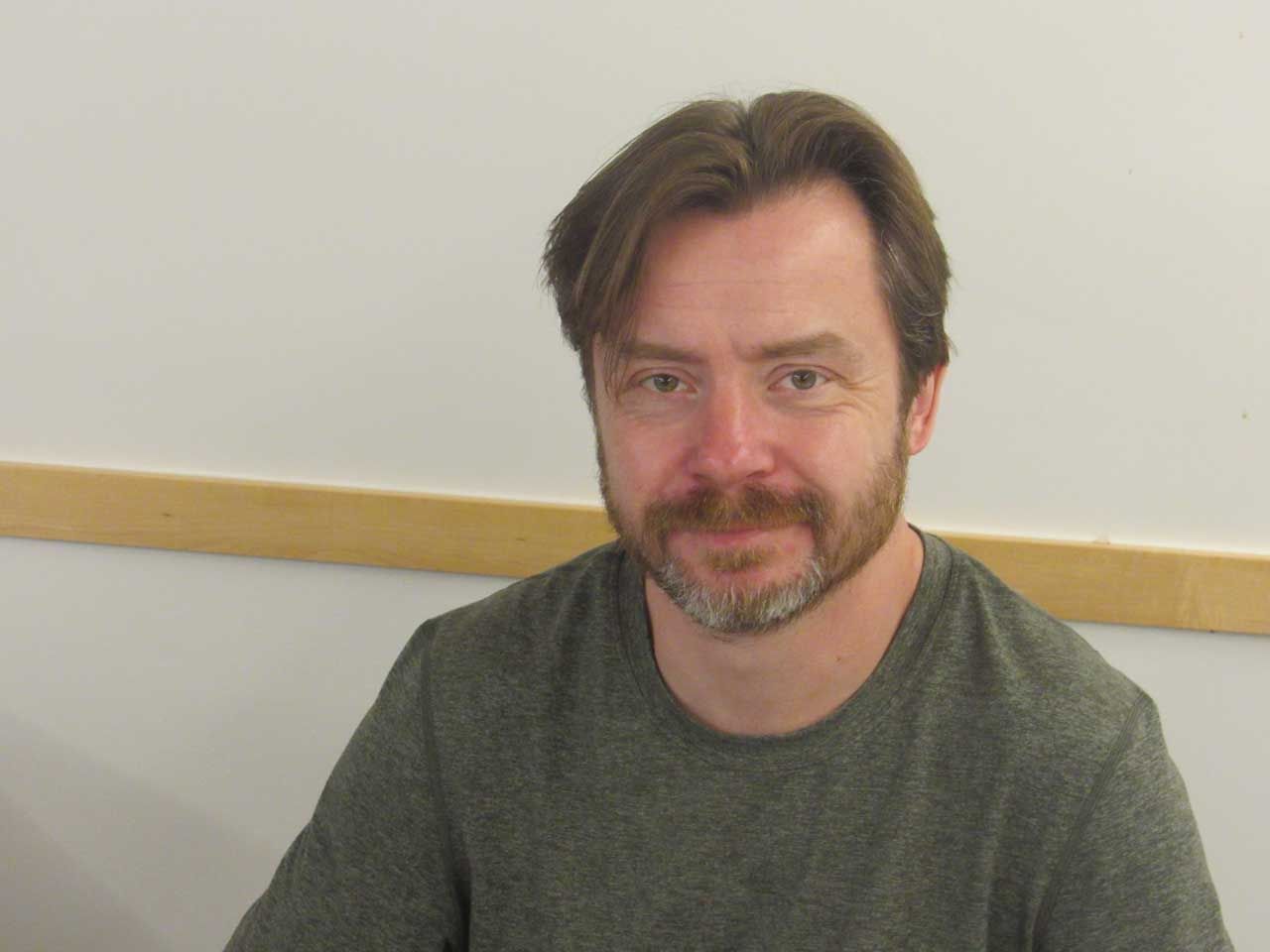 Man with brown hair and beard, wearing a gray shirt, smiling slightly, sitting indoors.