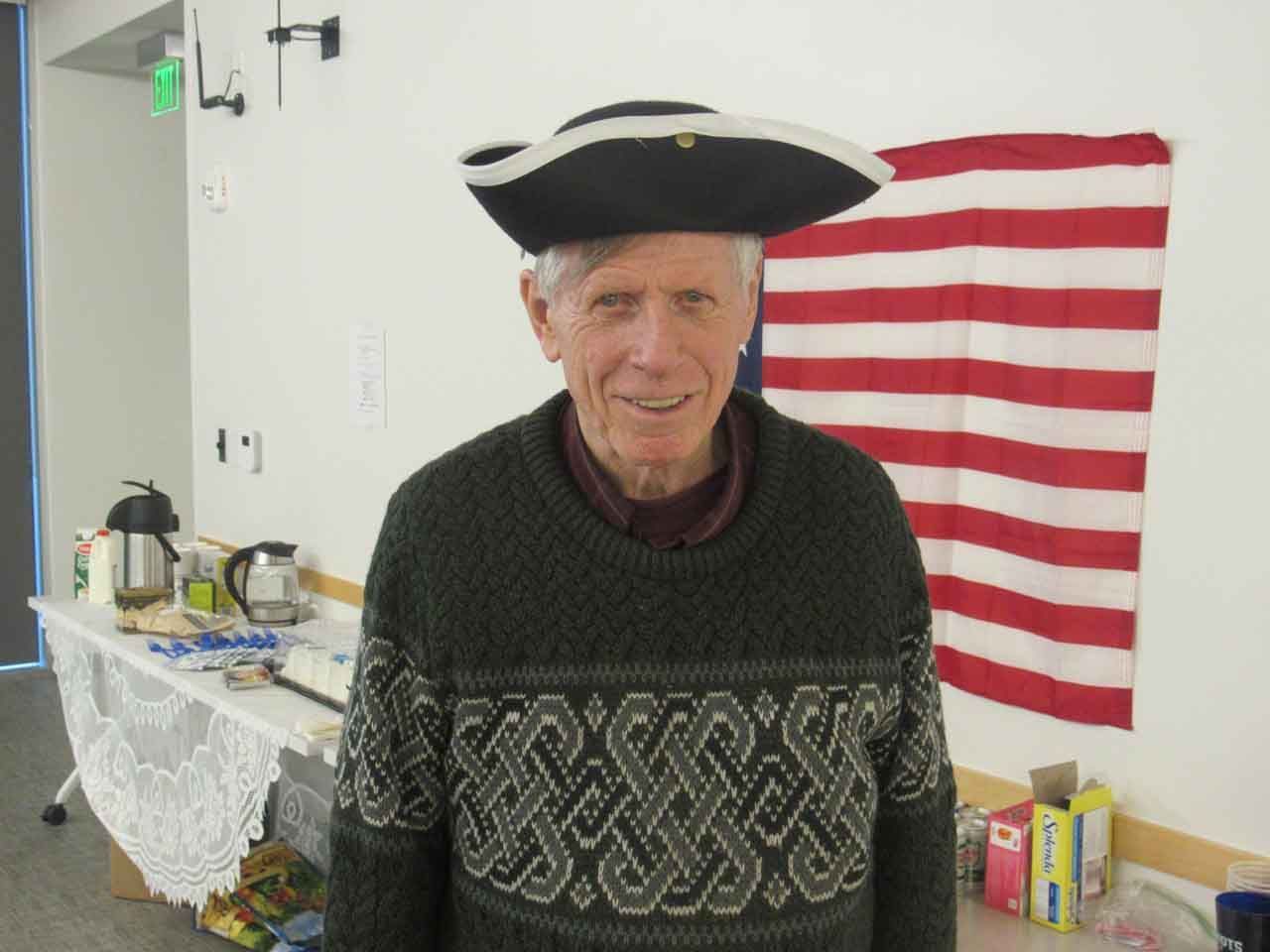 Man wearing a tricorn hat and patterned sweater smiles in front of an American flag.