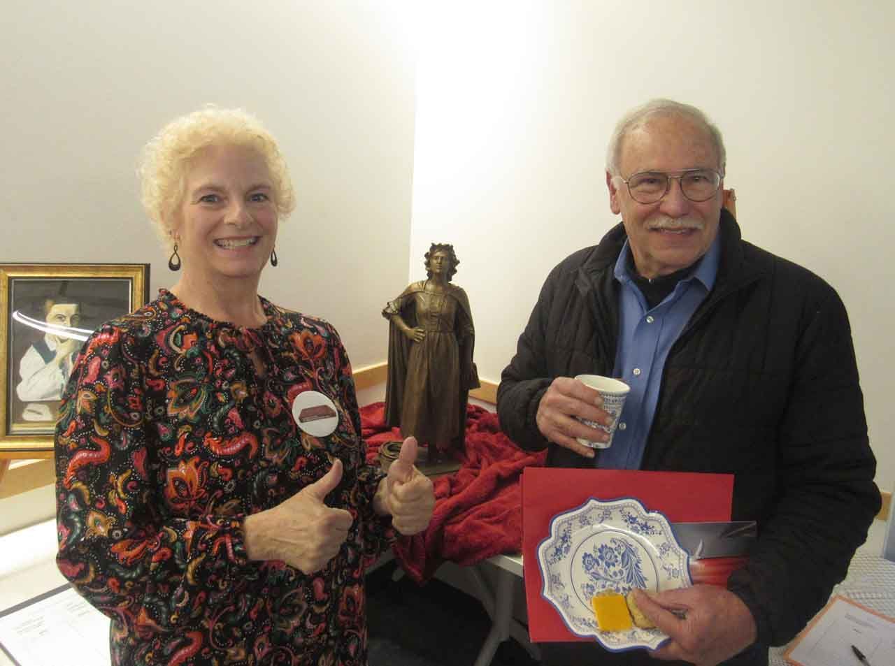 Woman gives thumbs up, man holds plate art, both by statue and painting.