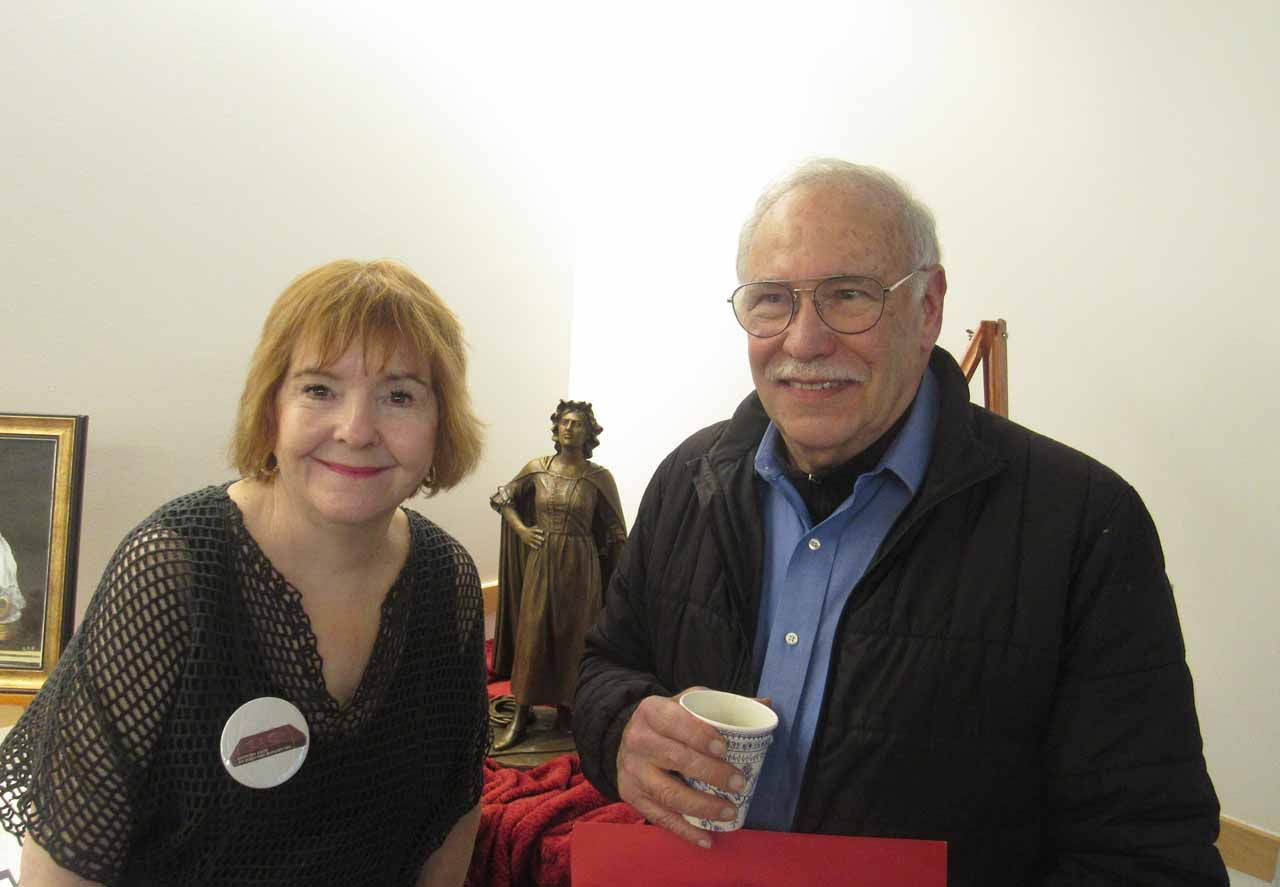 Woman and man smile, standing near a statue indoors. The man holds a cup.
