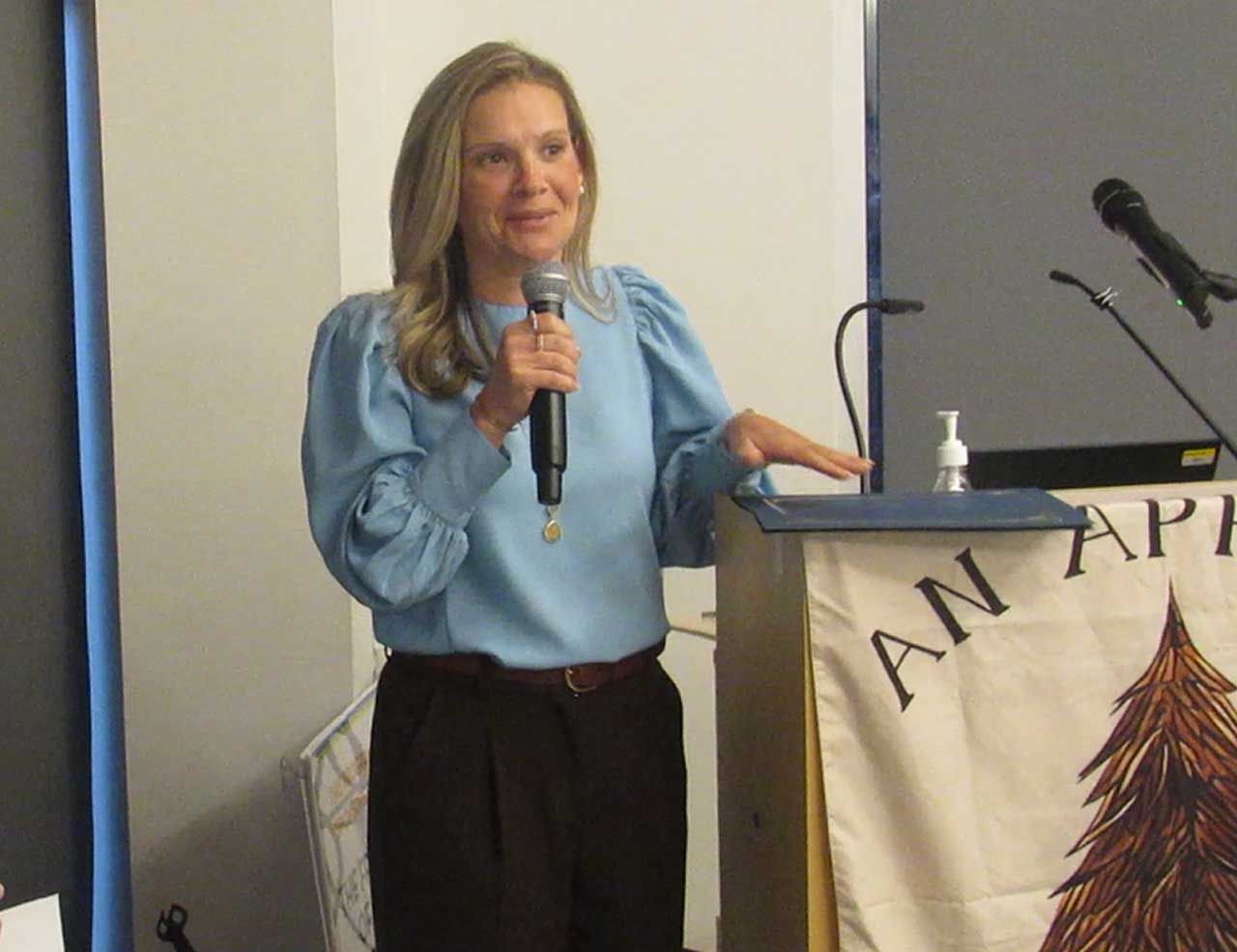 Woman speaking into a microphone at a podium with a banner that reads 