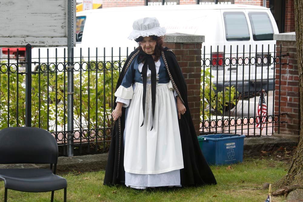 A woman in a white dress and black cape stands in front of a fence