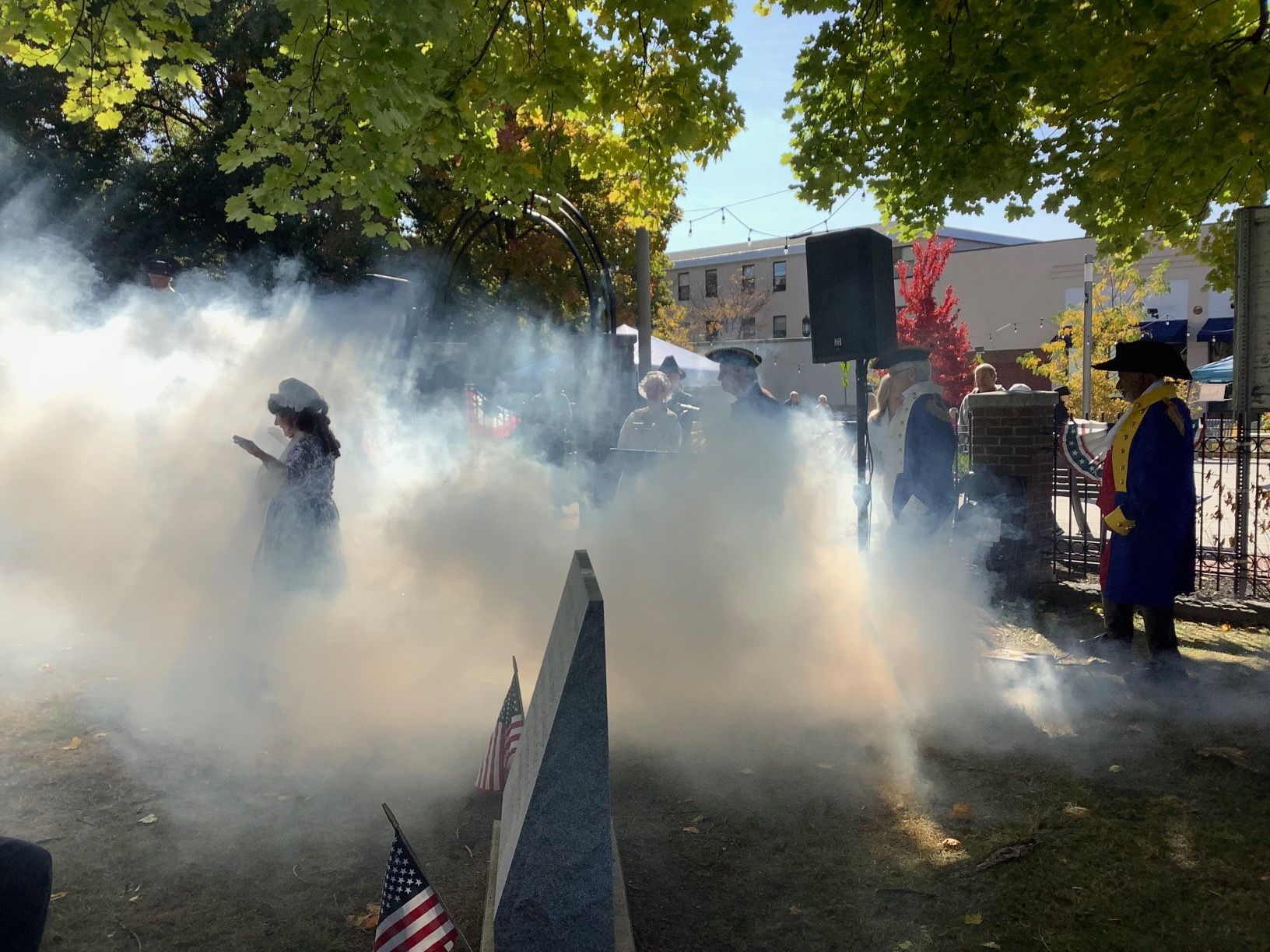 Ceremony with smoke. People in robes, American flags, and a monument. Outdoors, fall foliage.