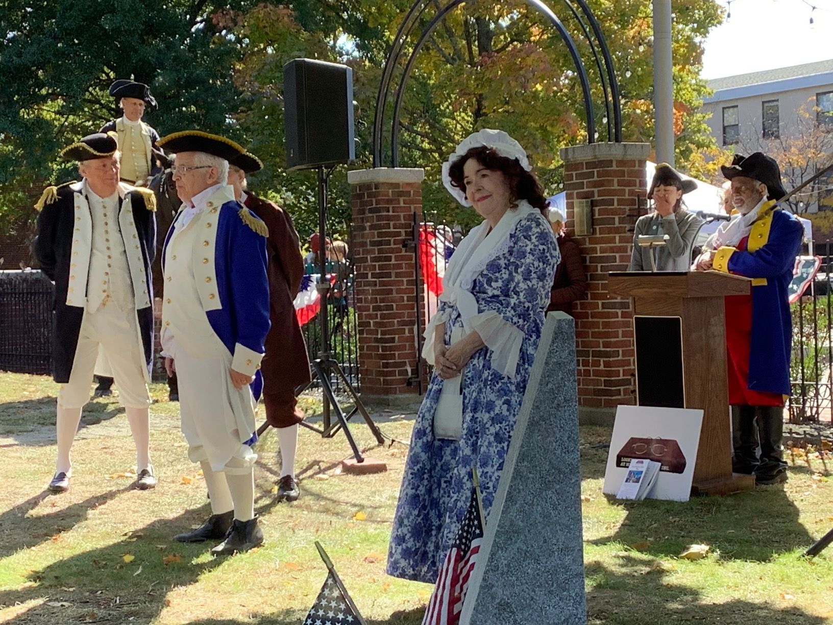 People in historical costumes at an outdoor event near a memorial, speaker.