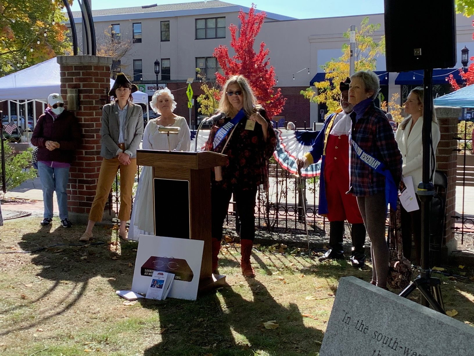 People at podium giving speeches, outdoors. Fall trees and buildings in background.