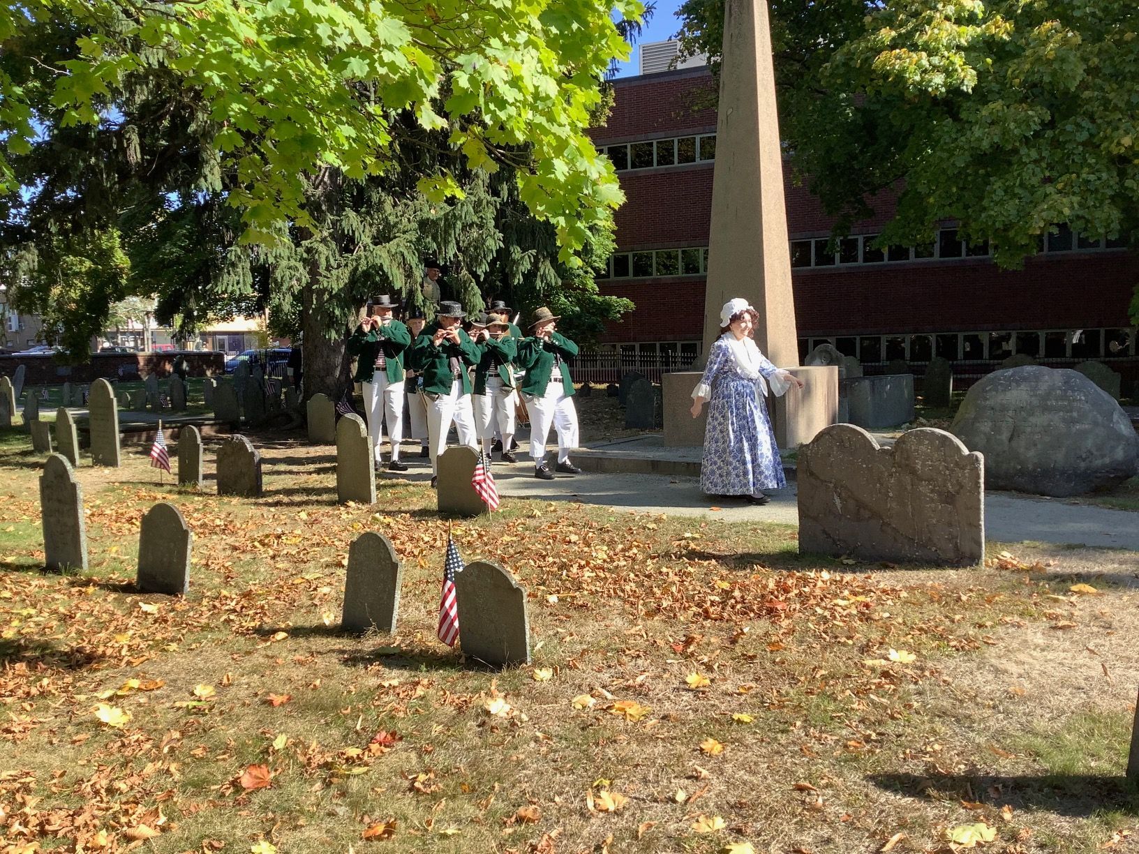 A group in colonial attire stands near an obelisk and gravestones in a cemetery, with soldiers in green.