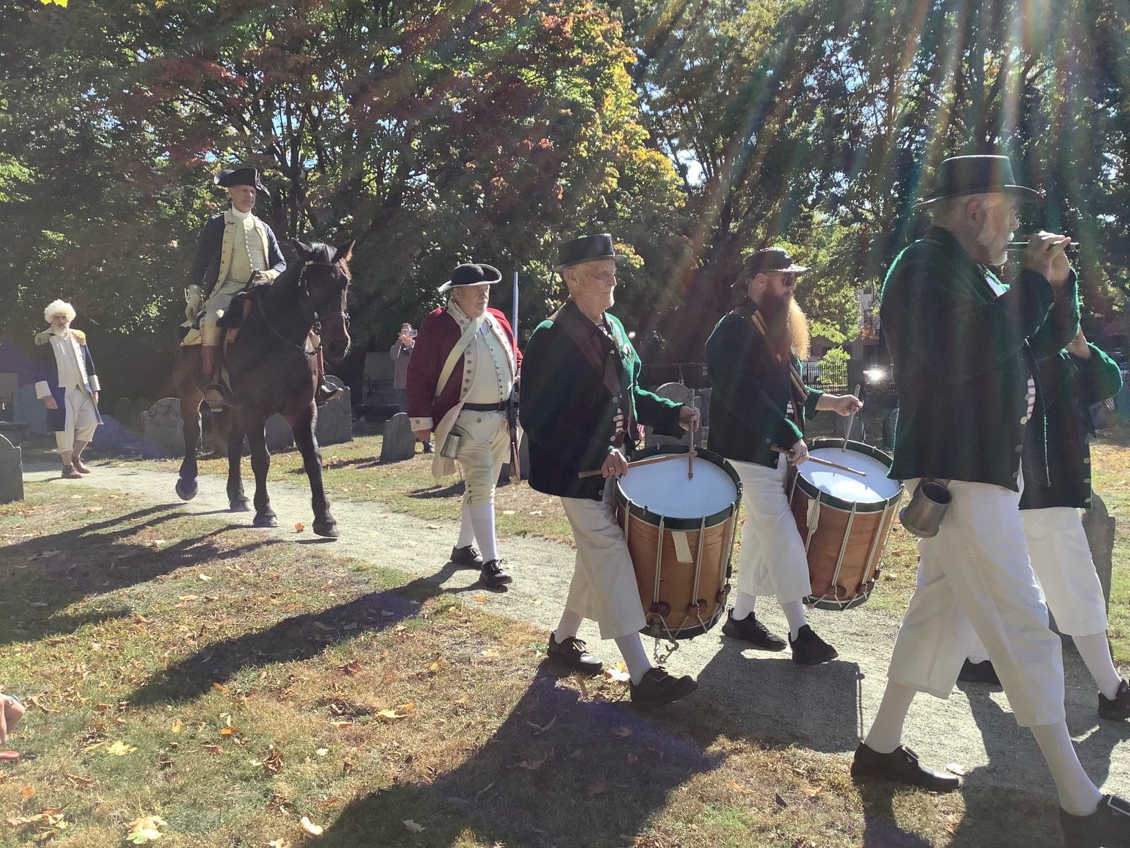 A historical reenactment: men in colonial garb march, some drumming, with a rider on horseback, outdoors.