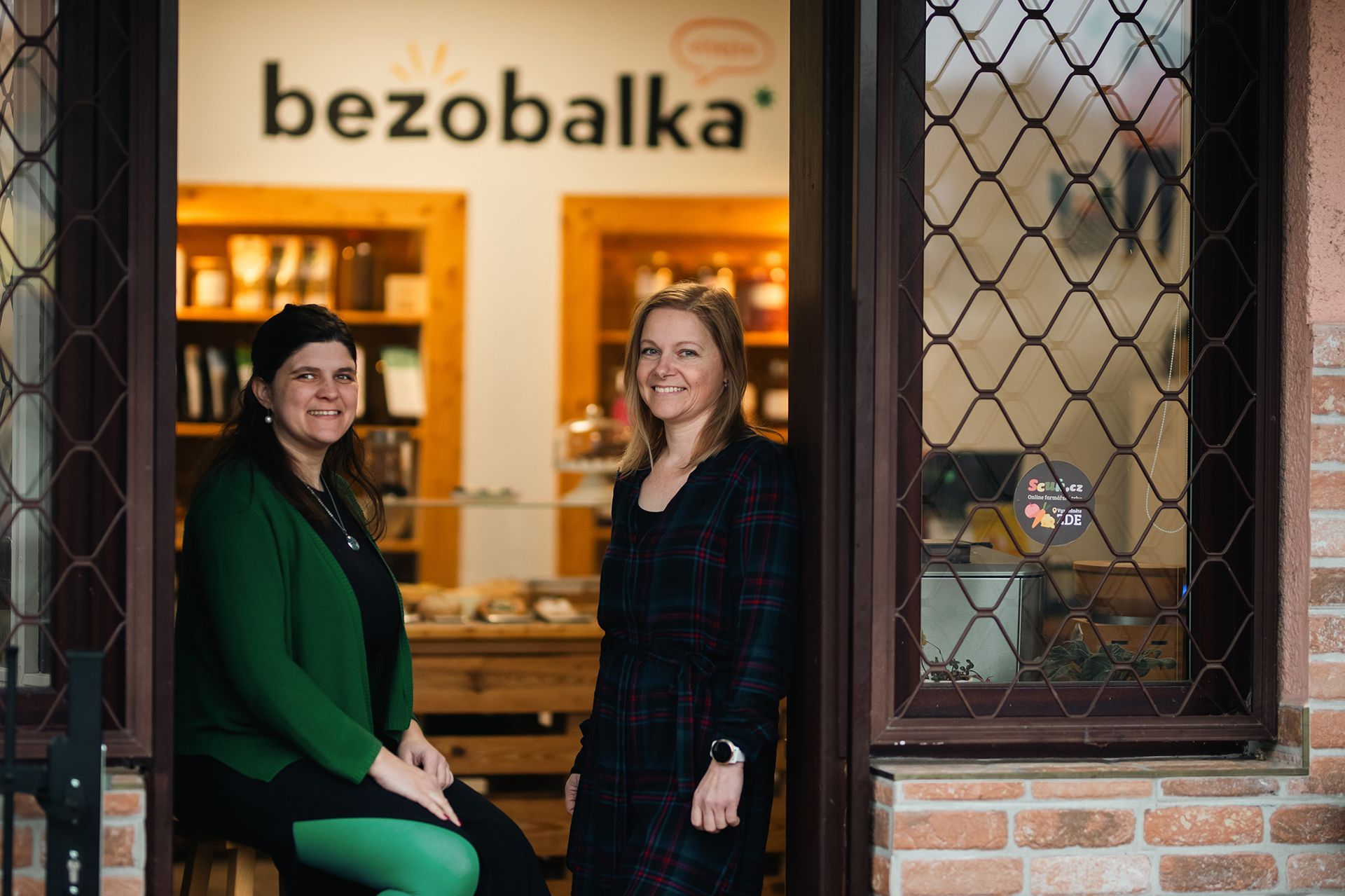 Dva lidé stojí ve dveřích obchodu s názvem „bezobalka“ a uvnitř je teplé světlo.