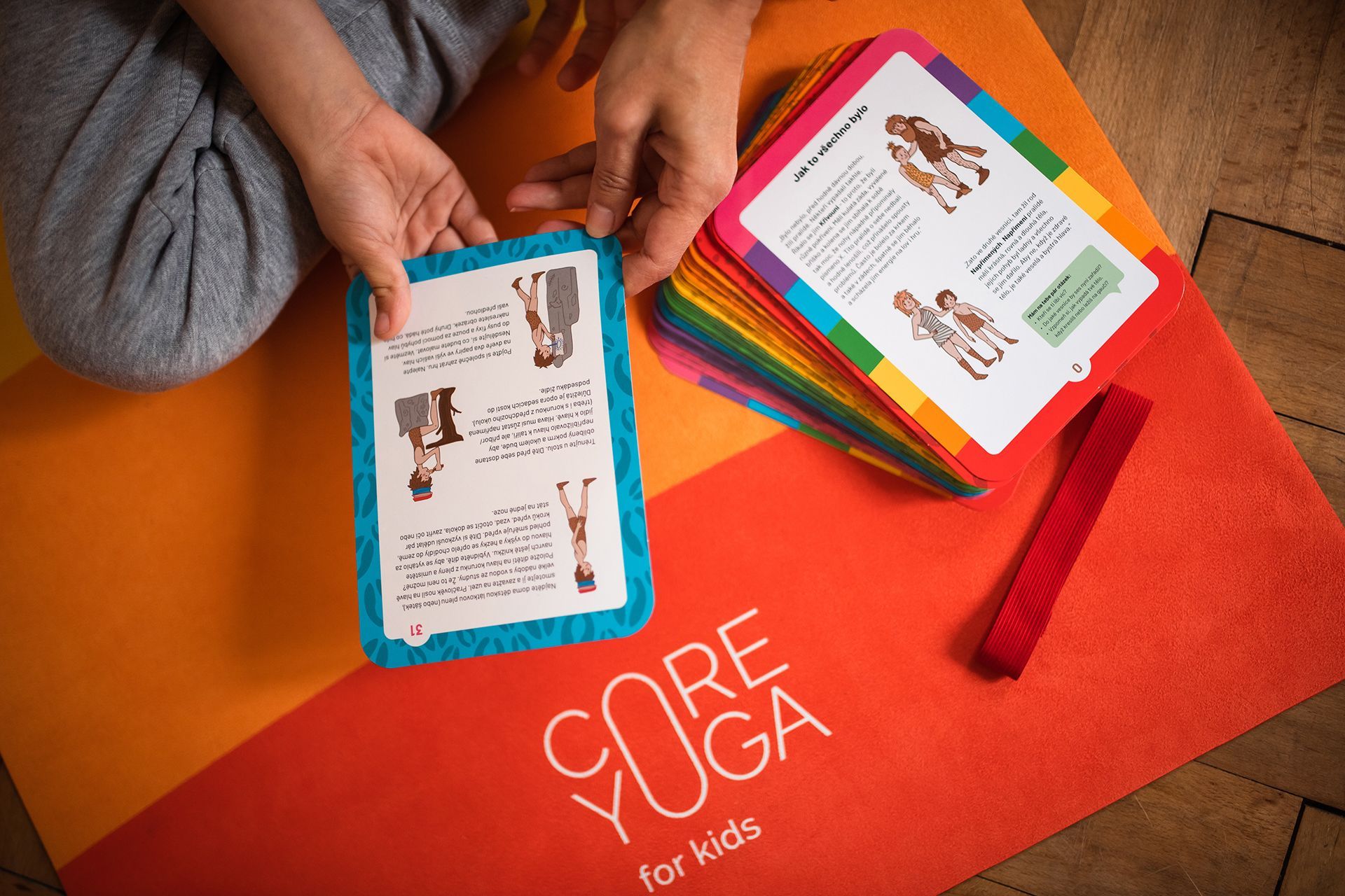 Ruce držící barevné dětské kartičky na oranžové podložce „CORE Yoga for Kids“