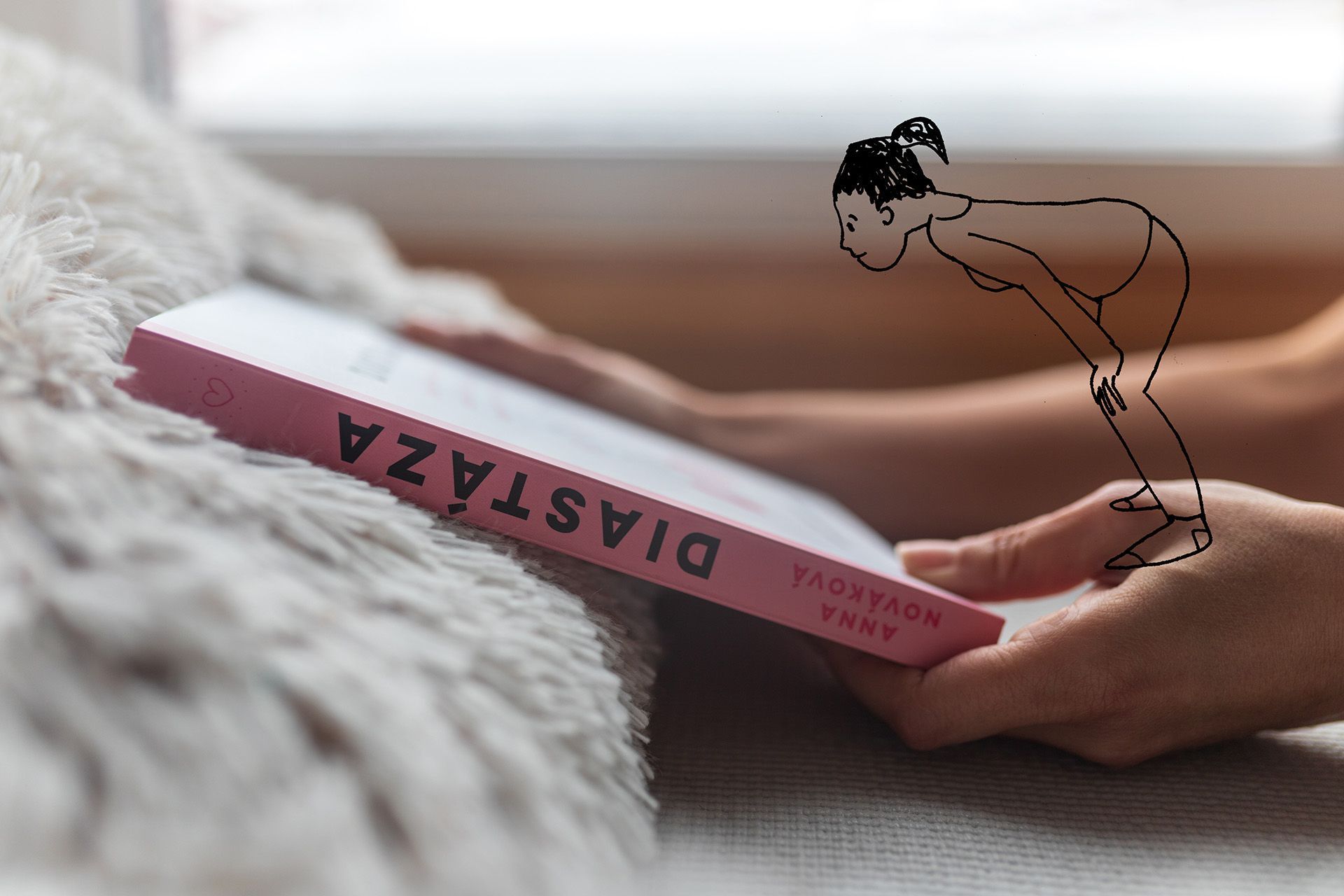 Růžová kniha položená na posteli vedle nadýchané bílé deky s kresbou osoby naklánějící se nad ni