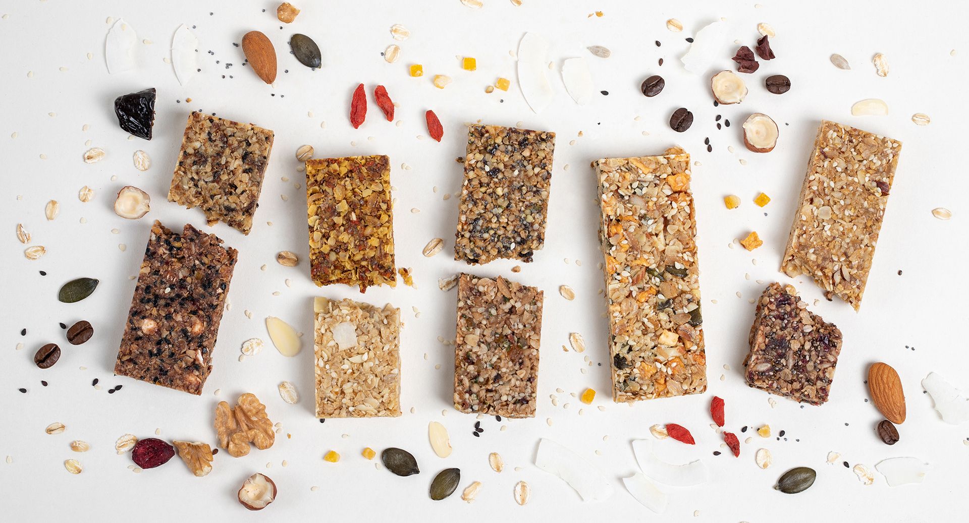 Granola tyčinky a roztroušené ořechy na bílém pozadí, uspořádané v řadě s drobkami kolem nich