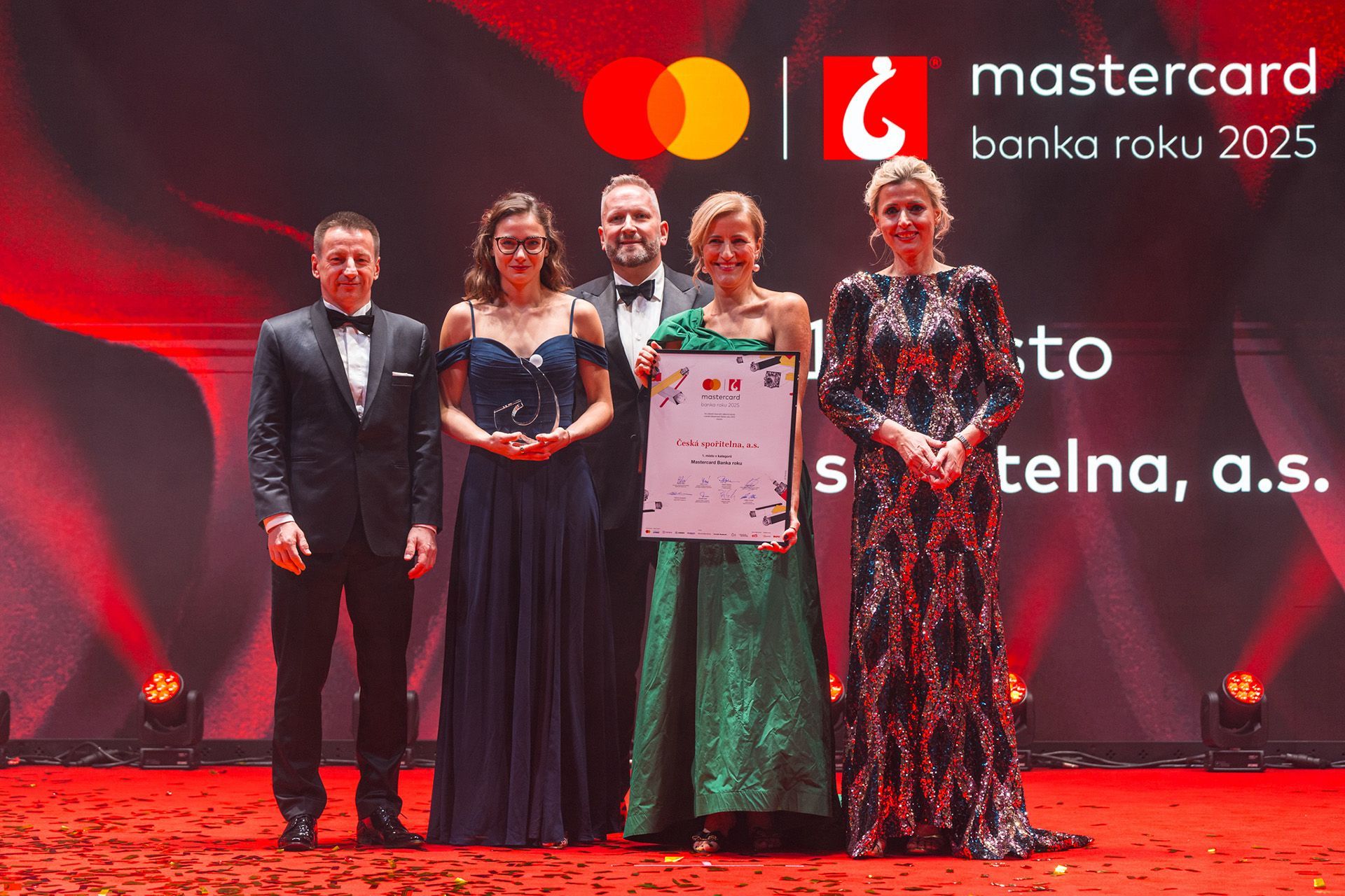 Čtyři lidé na červeně osvětleném pódiu pro předávání cen držící certifikát, v pozadí loga Mastercard a banka roku 2025