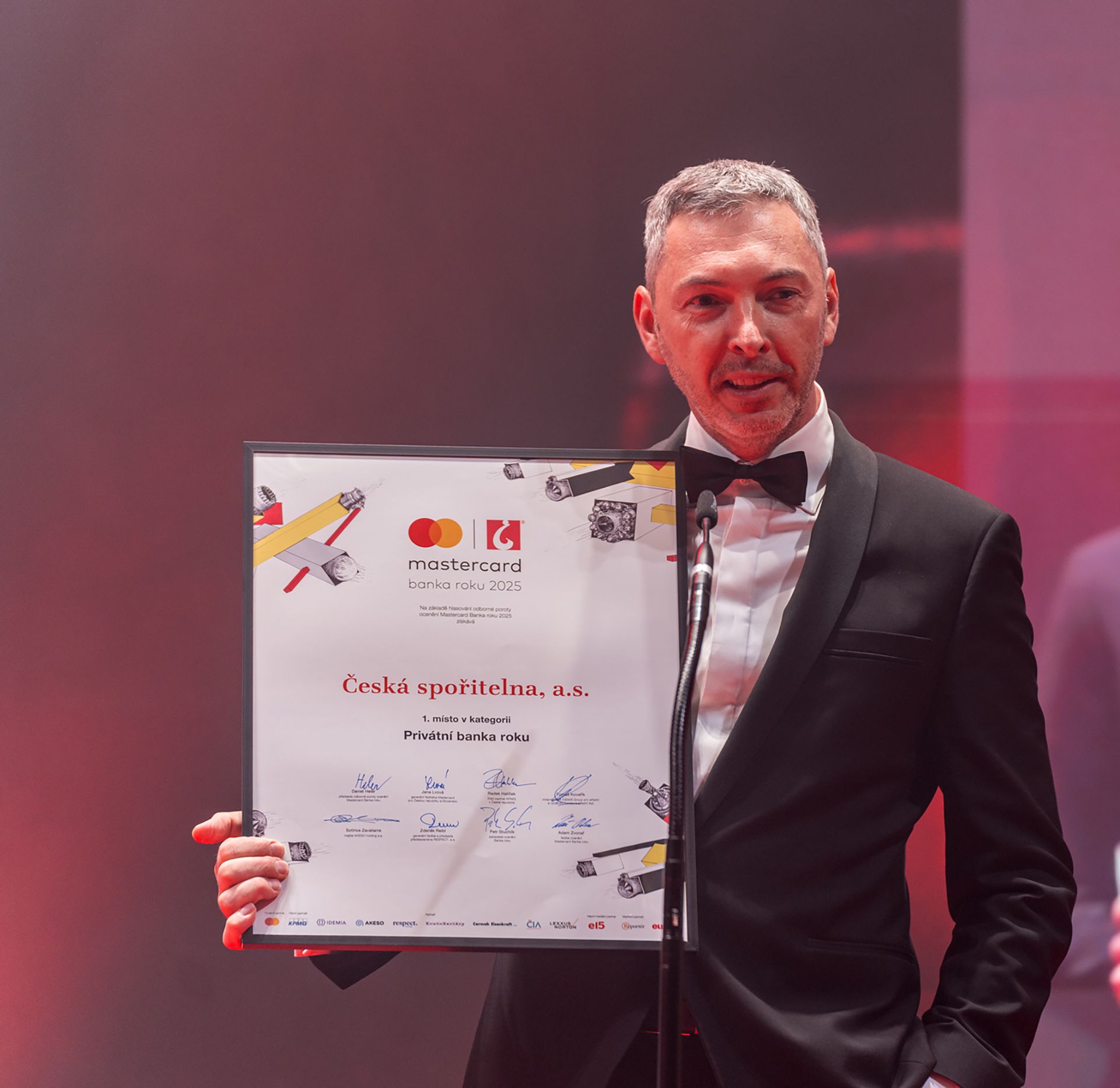 Muž ve smokingu držící certifikát na pódiu vedle mikrofonního stojanu, pod růžovým osvětlením.