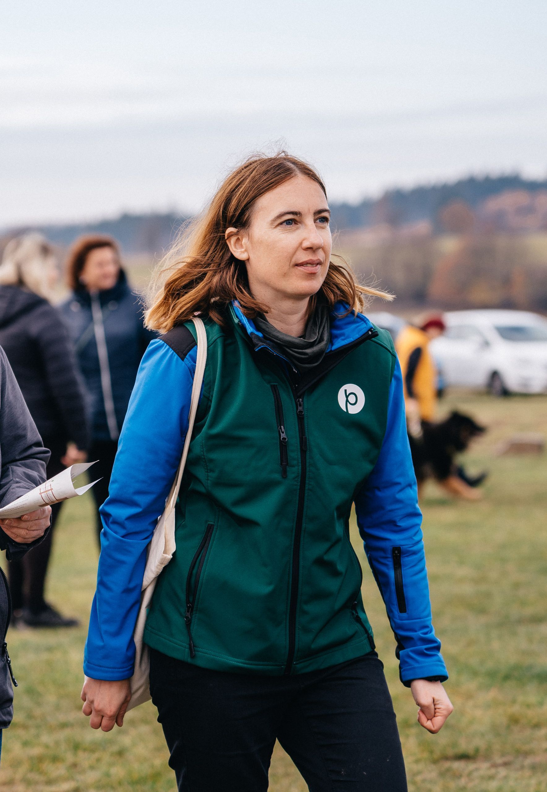 Žena v zelené vestě a modré bundě prochází se venku po travnatém poli s ostatními v pozadí